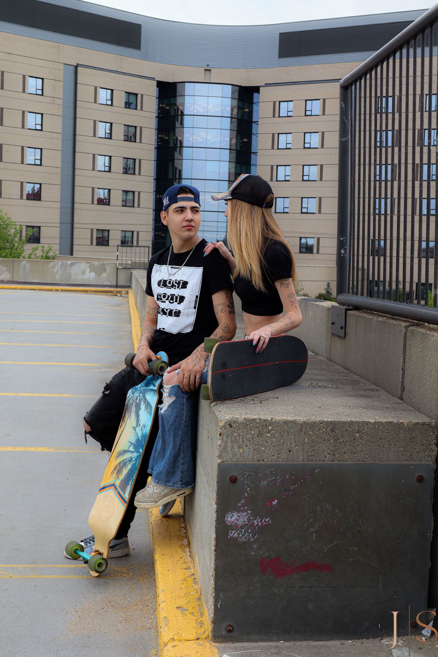 Two skateboarders sit casually on a concrete barrier in an urban parking lot with a modern building in the background.
