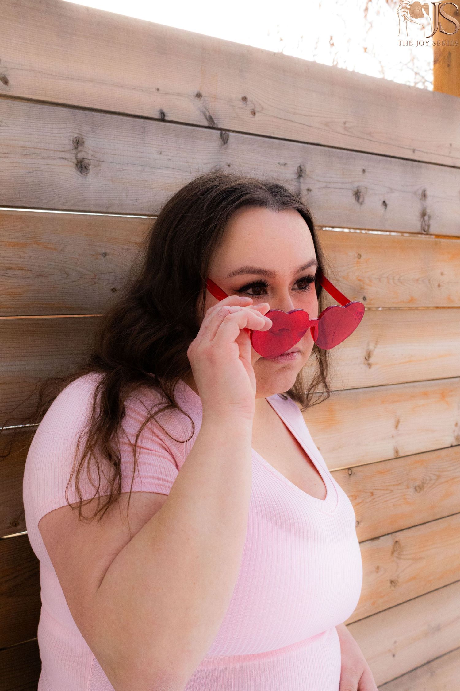 Fashion model in pink top poses playfully with heart-shaped sunglasses against wooden wall backdrop.