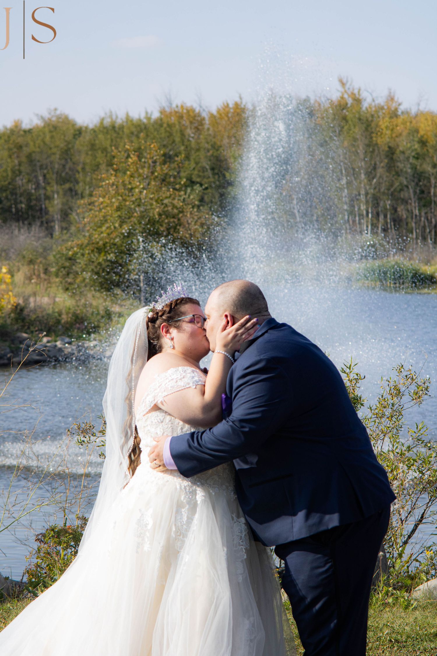 A romantic wedding couple shares a kiss by a lake with a water fountain spray creating a magical backdrop.