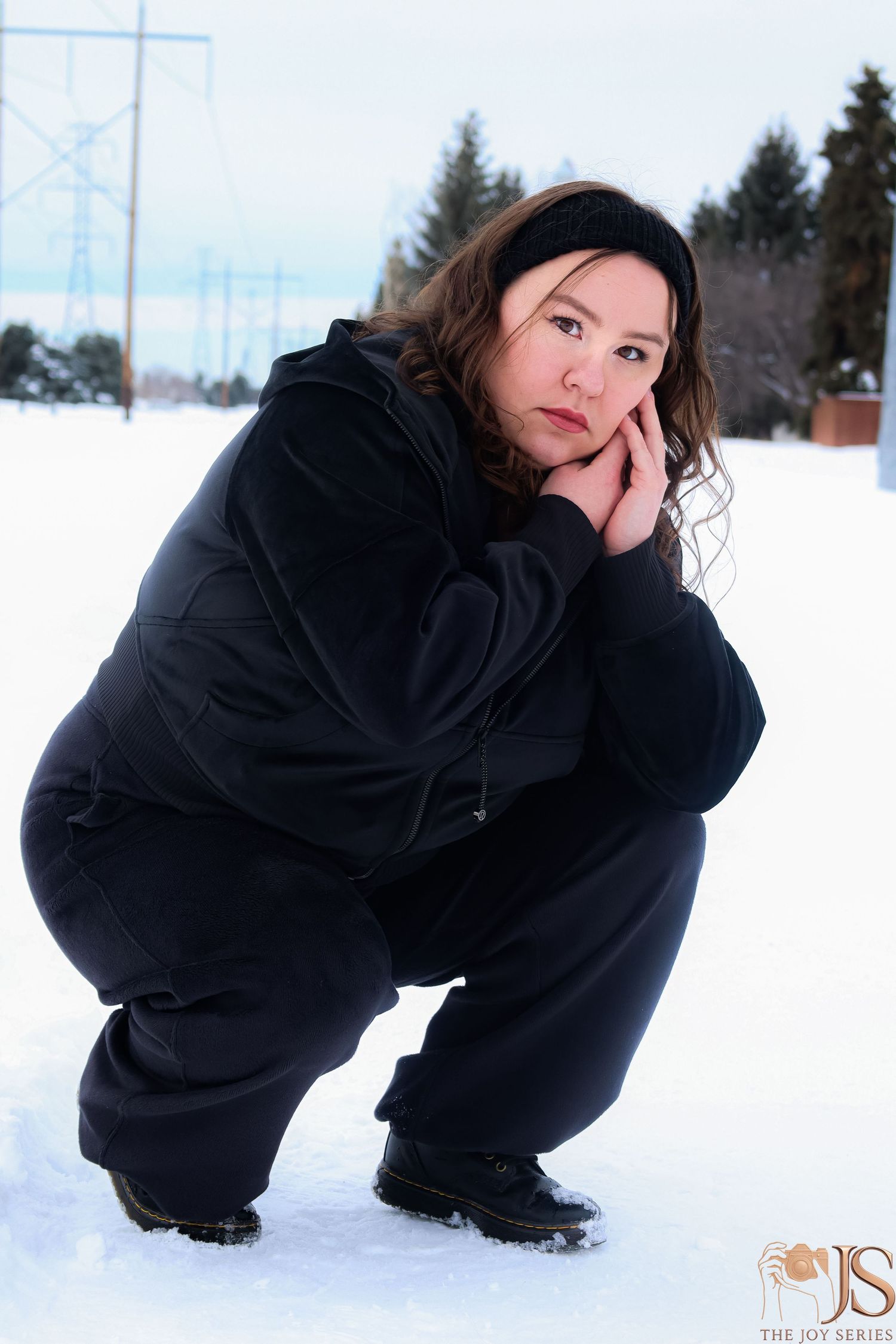 Person in black winter clothing crouching playfully in snowy landscape with power lines visible in background.