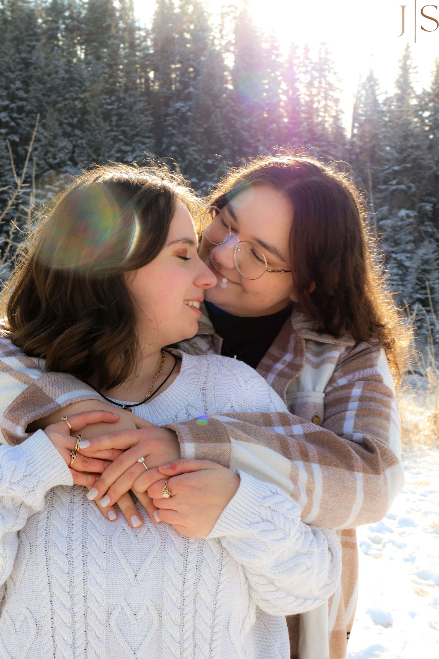 A romantic winter photoshoot showing a couple in white sweaters embracing against a snowy forest backdrop at sunset.