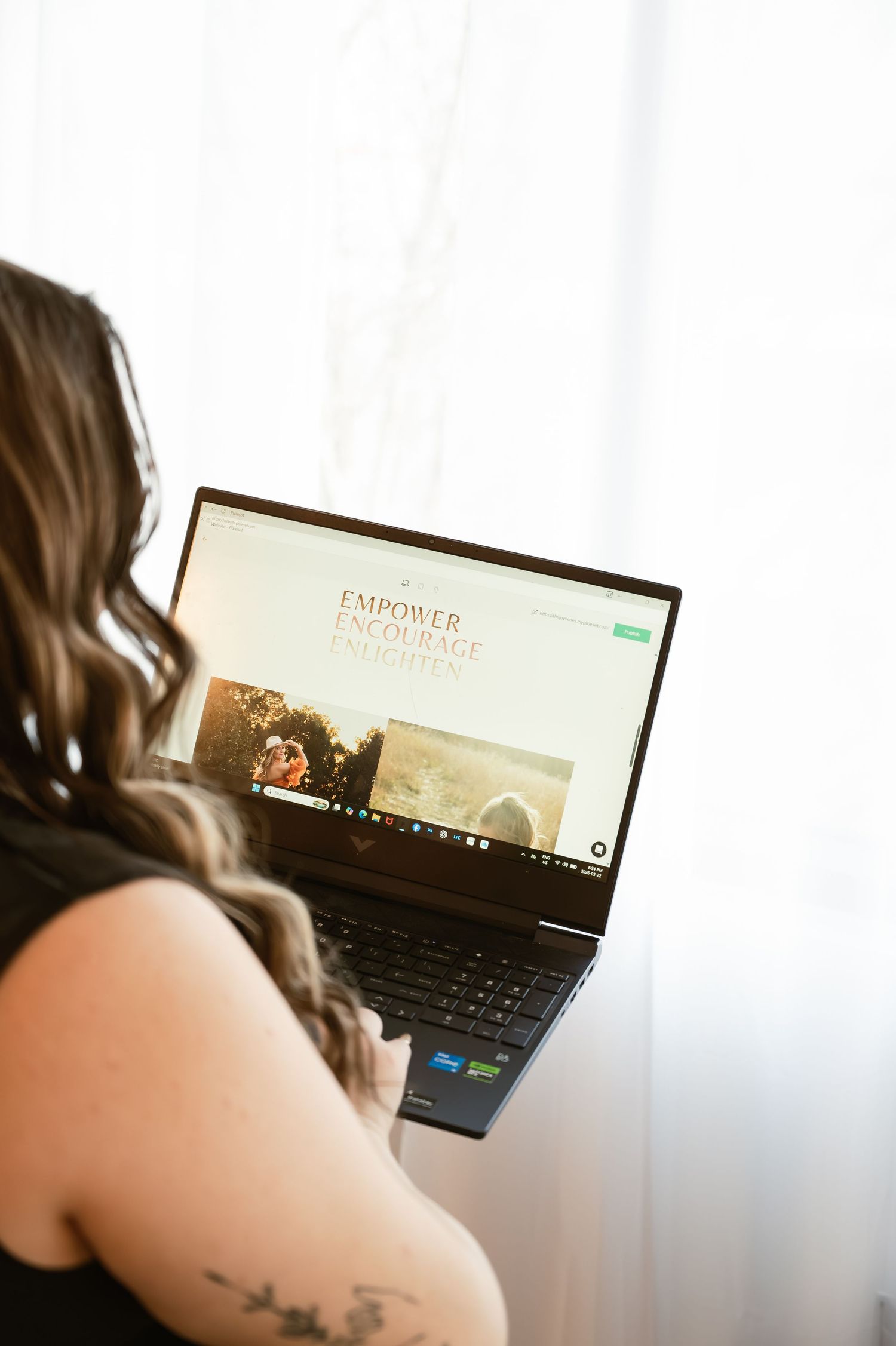 Woman with tattoo viewing an Empower Encourage website on an HP laptop near a bright window.