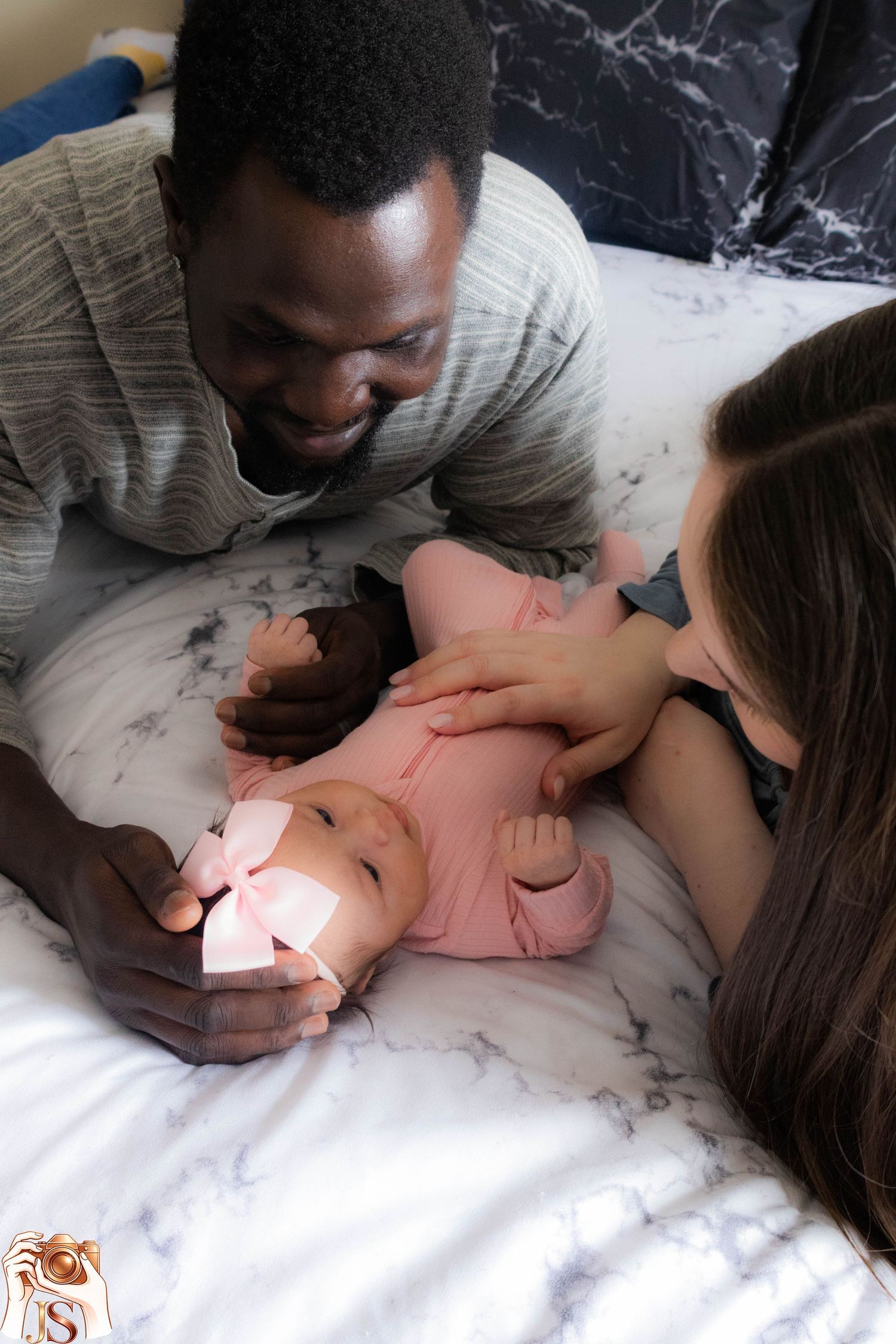 A baby in a pink outfit lies on white bedding while two adults lean in close and share a tender moment together.