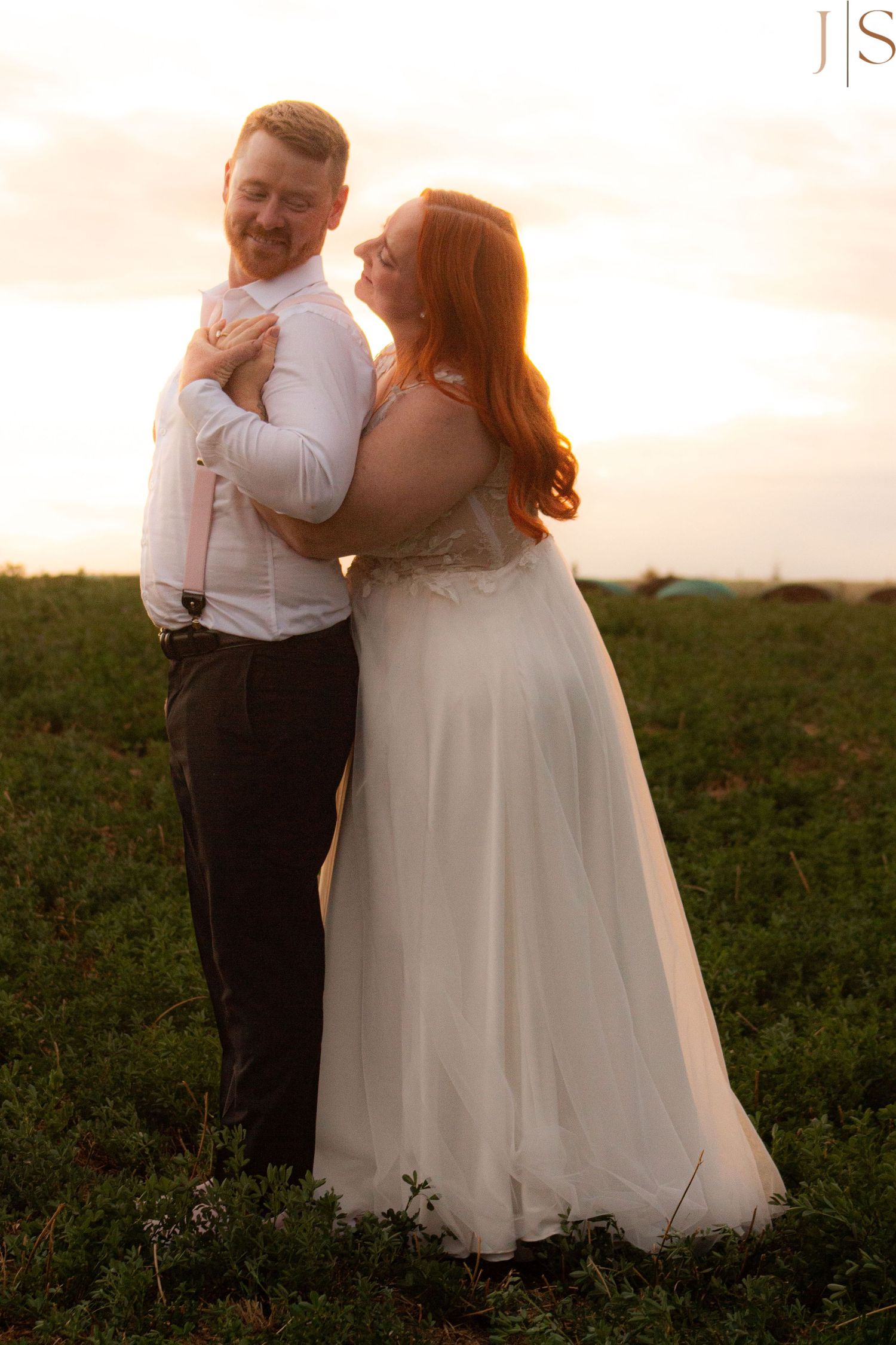 Newlyweds embrace in a field at sunset, with the bride wearing a flowing white gown.