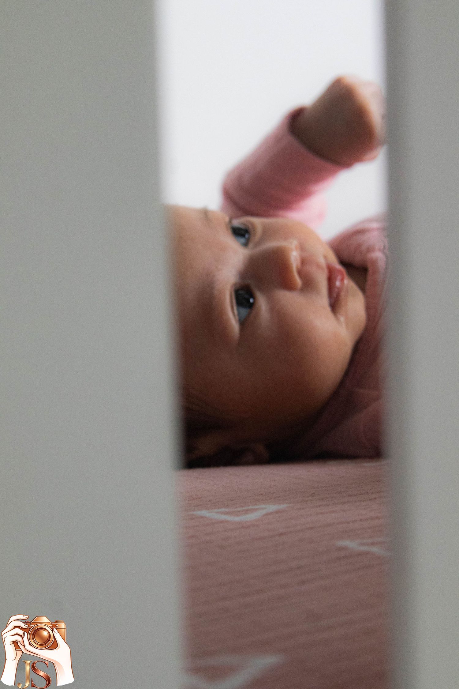 A baby lying on pink bedding viewed through a gap, with soft lighting creating an intimate nursery scene.