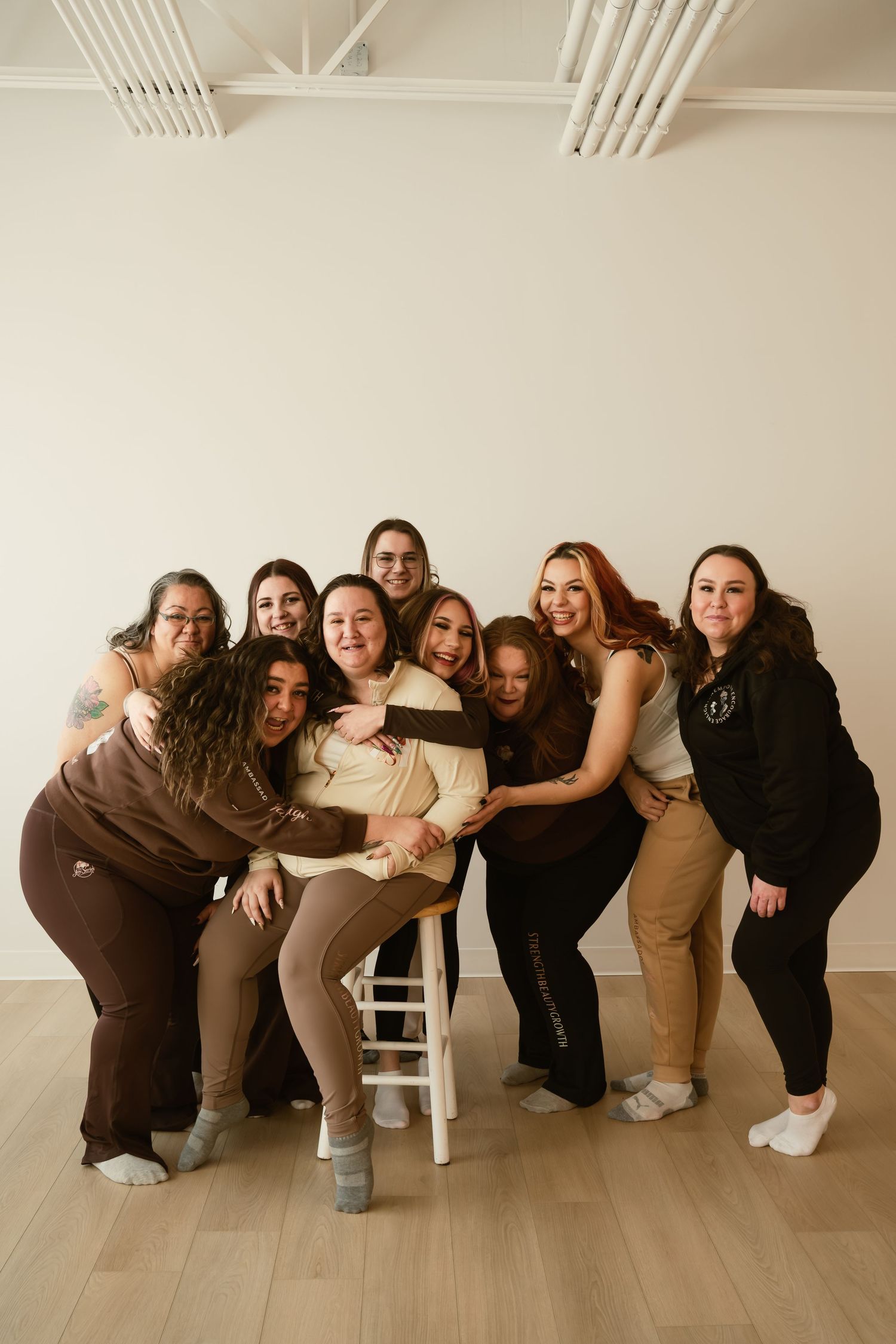 Group of nine women in brown and neutral tones posing together, smiling and hugging in a bright studio space.