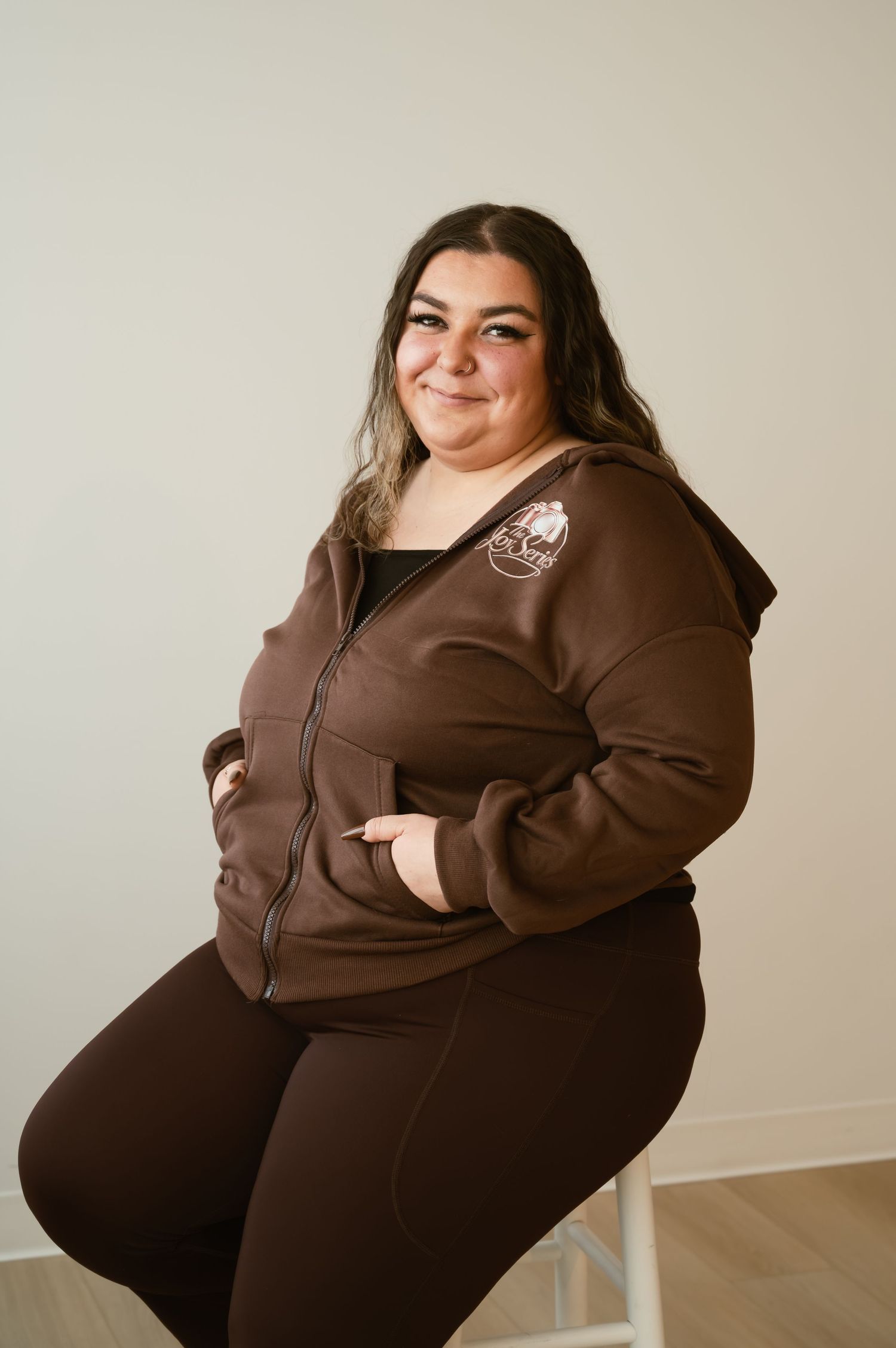 Woman in brown zip-up hoodie and matching pants, smiling confidently while seated against a neutral background.