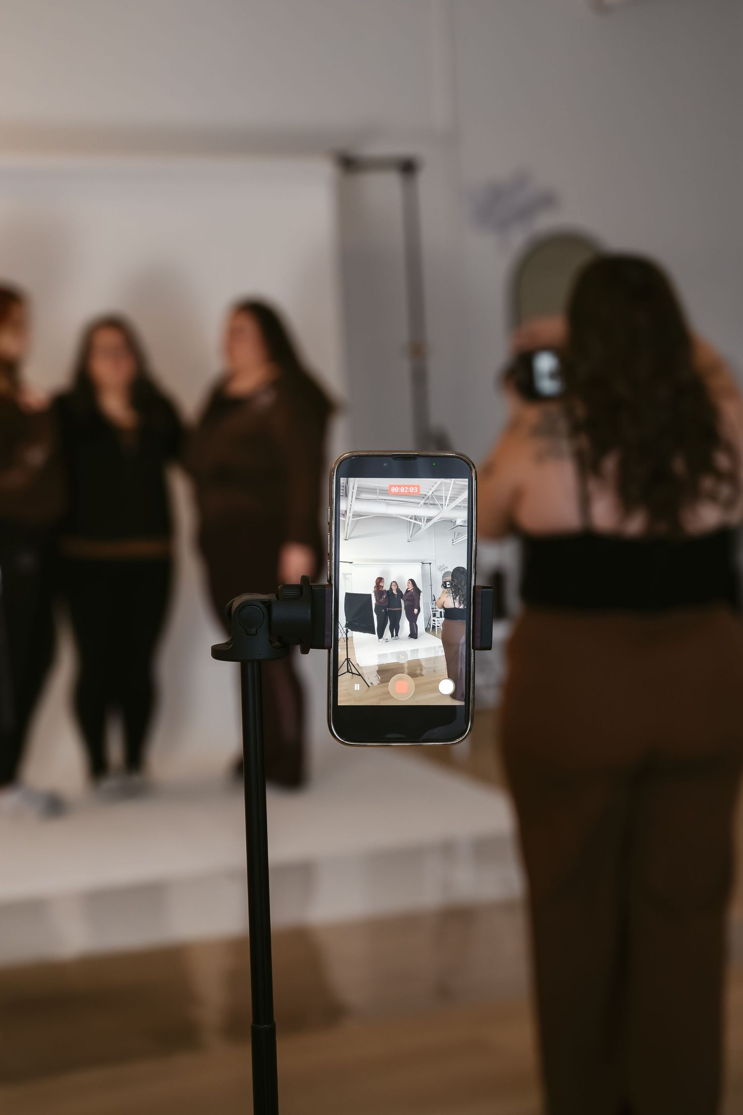 A smartphone on a tripod captures two women posing during an indoor photo shoot, with others visible in the blurred background.