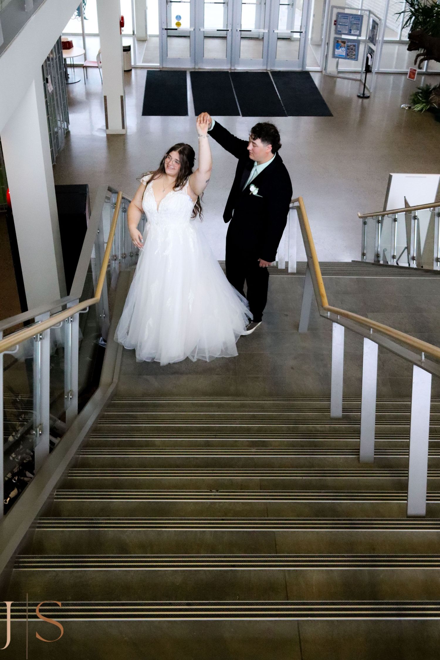 A couple in formal wedding attire pose on a modern staircase with glass walls and handrails in an open building atrium.