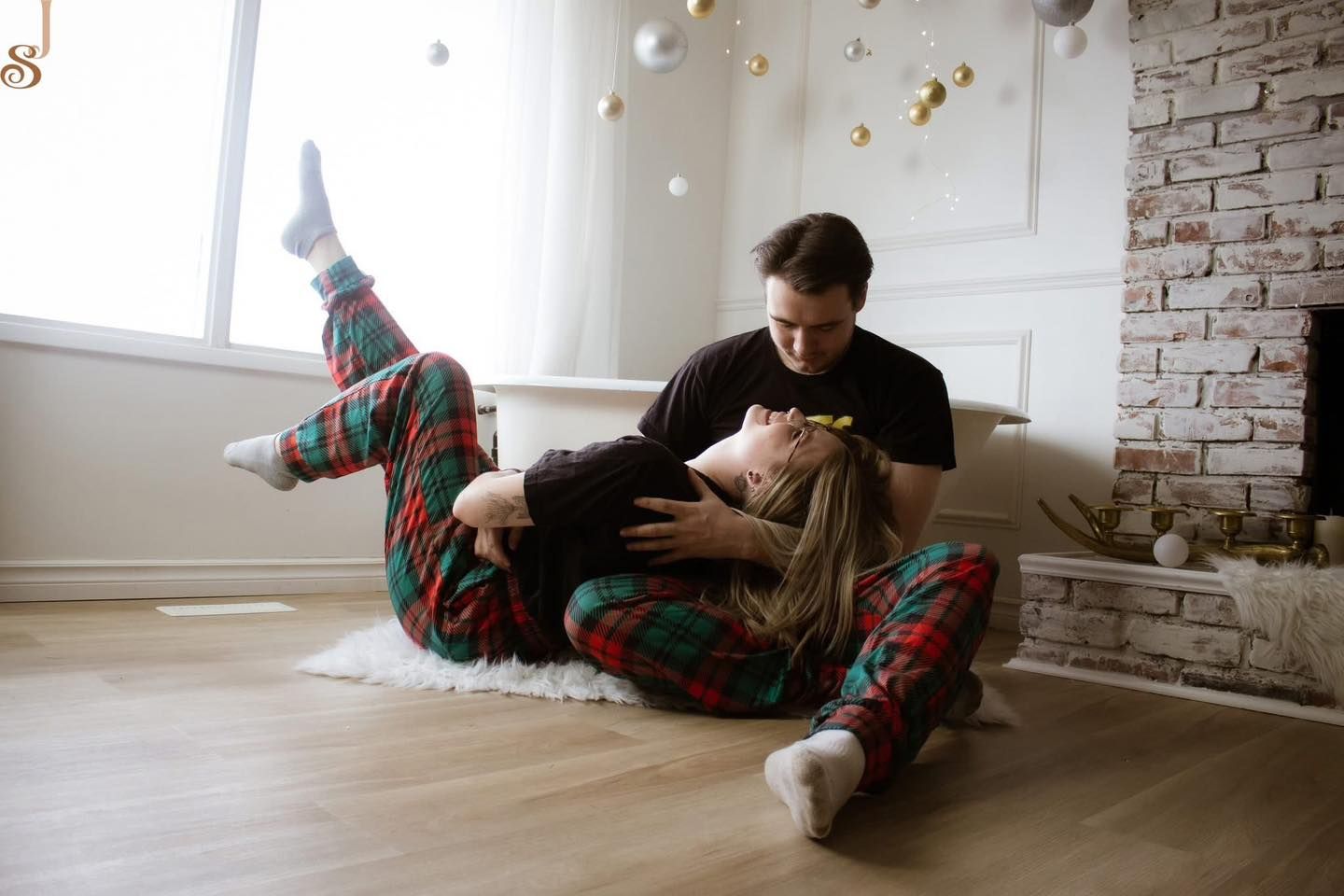 Two people in cozy holiday pajamas share a tender moment while sitting on a hardwood floor near an exposed brick wall.