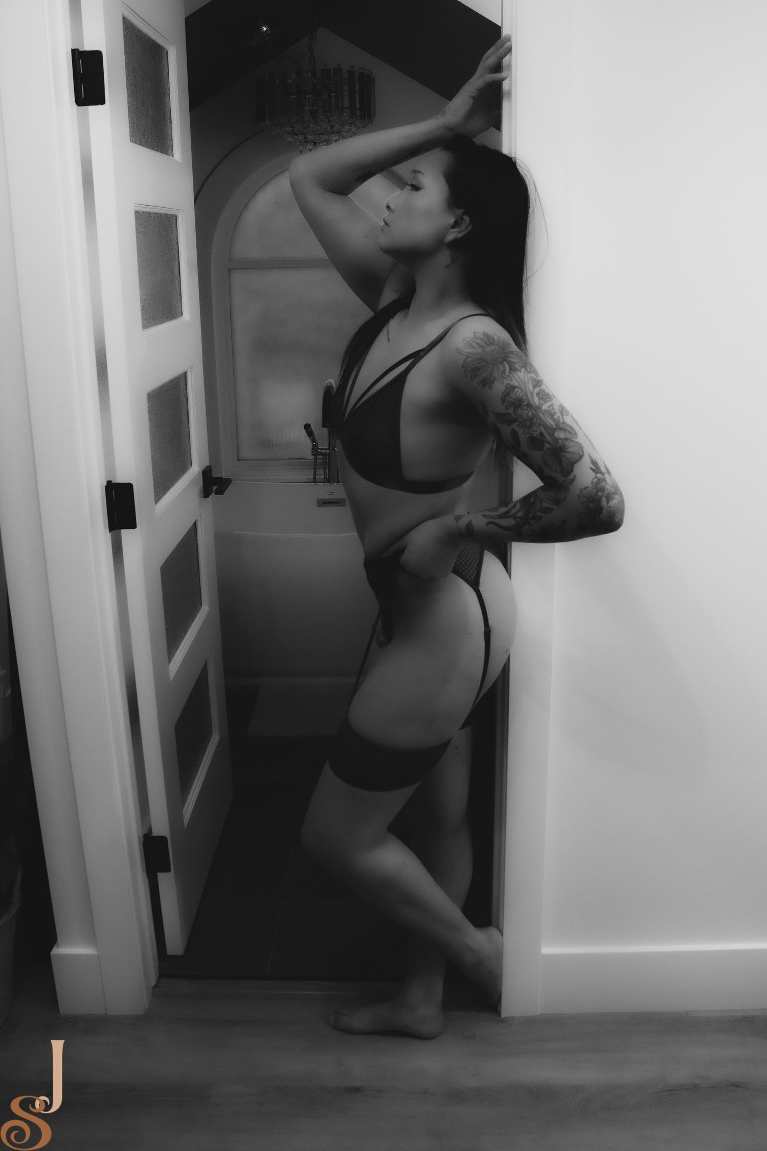 A black and white artistic photo showing a person in lingerie with tattoos posing against a doorway.