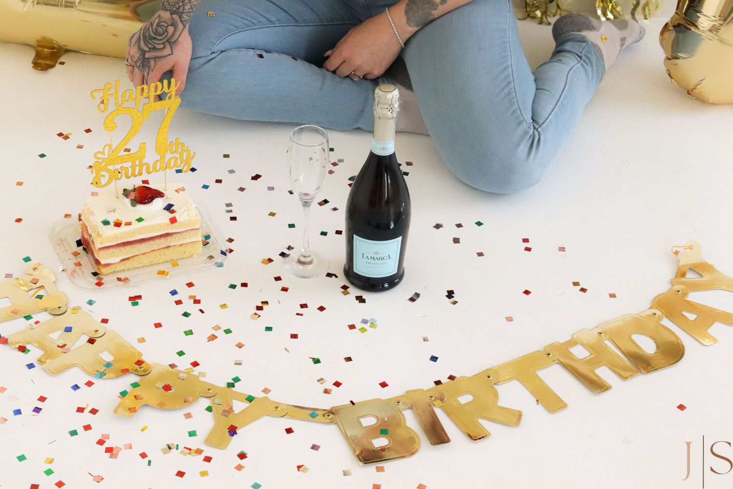 A birthday celebration setup with champagne bottle, confetti, and gold 'BIRTHDAY' banner on white floor.