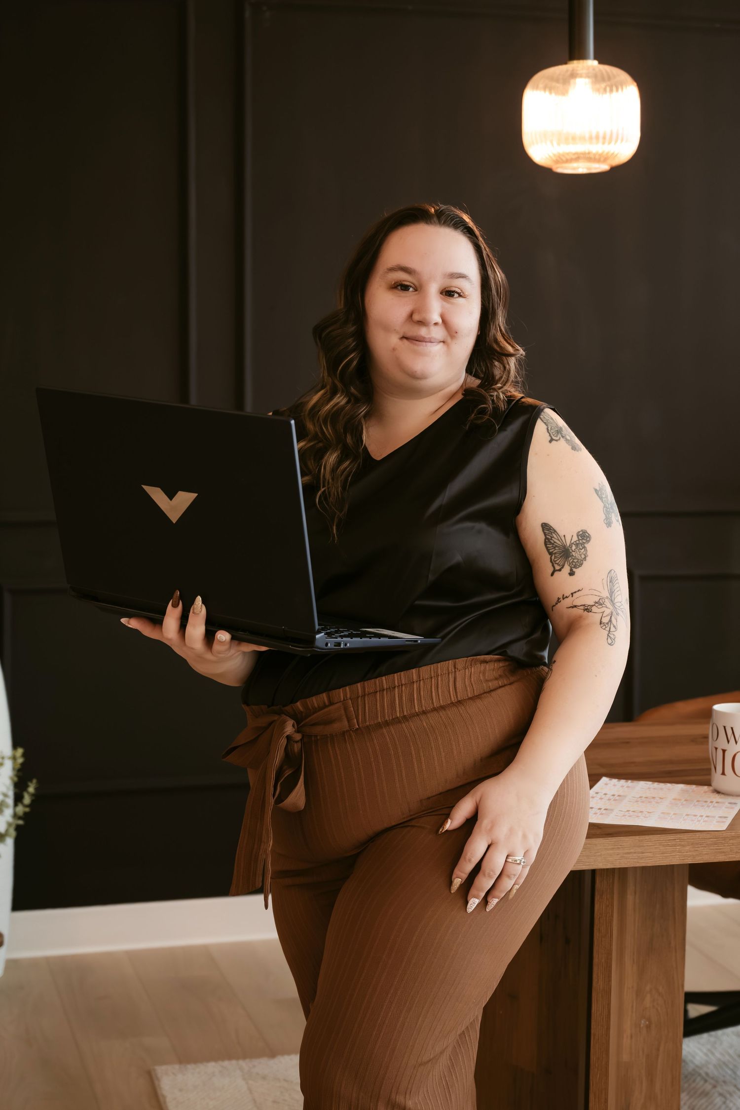 Woman holding HP laptop, wearing black top and brown pants, with tattoos, posing in modern dark-walled office space.