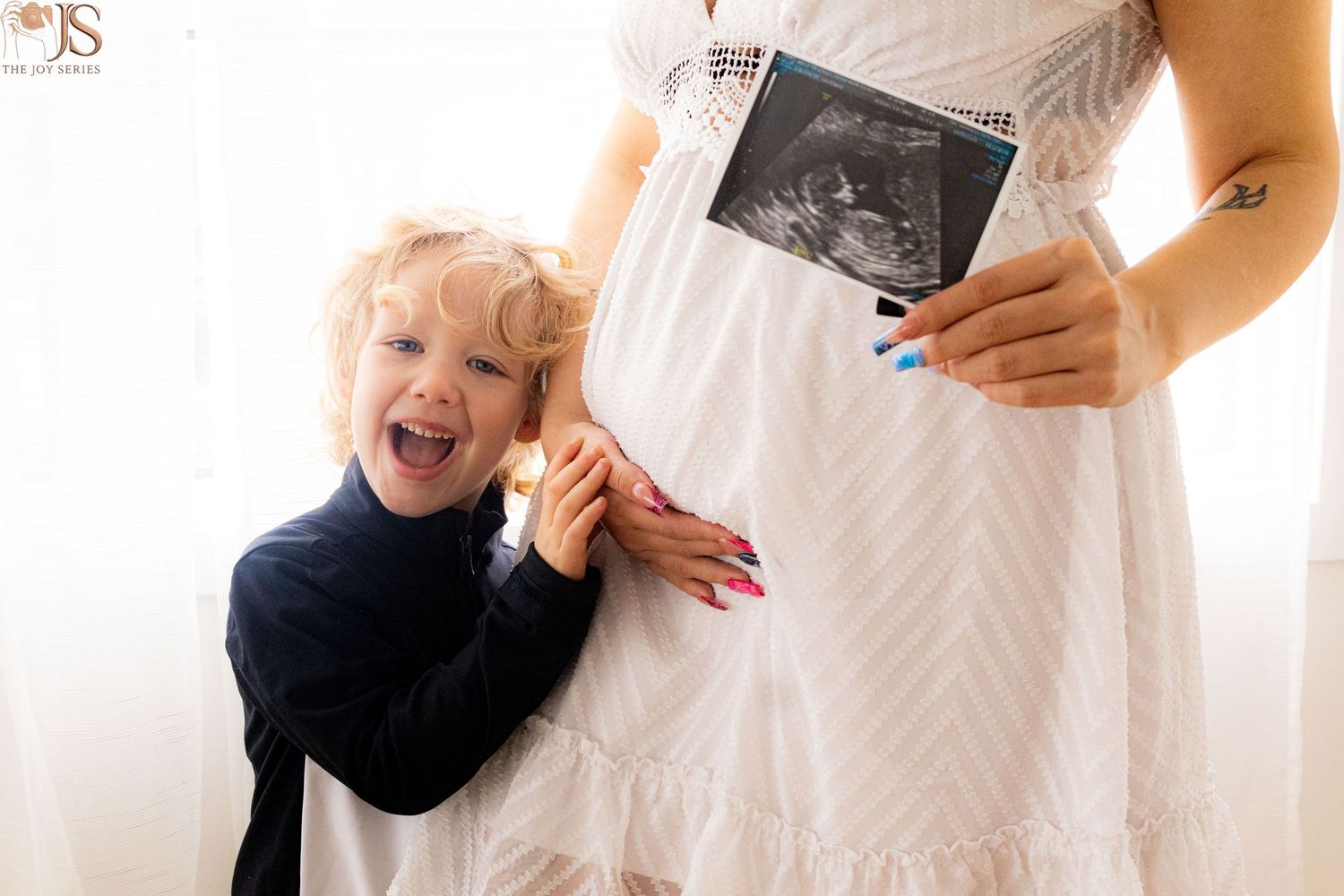 Excited toddler boy smiles beside pregnant mother in white dress holding ultrasound photo during maternity photoshoot.