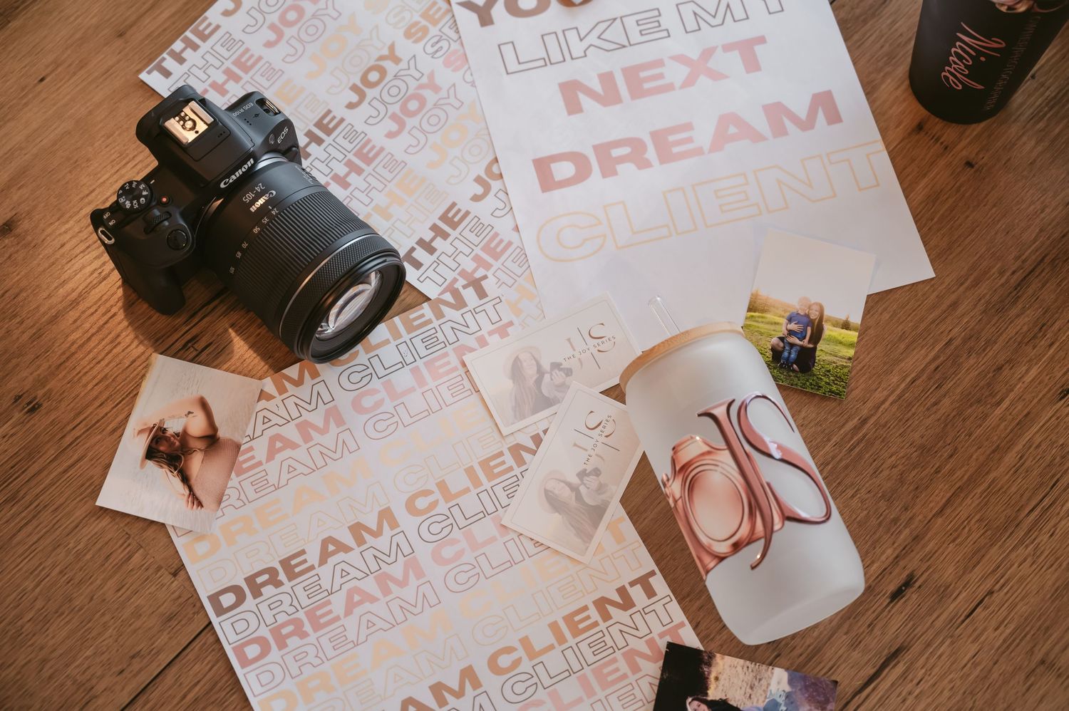 Camera, dream client posters, photos, and branded merchandise arranged on a wooden floor for a photography business flatlay.