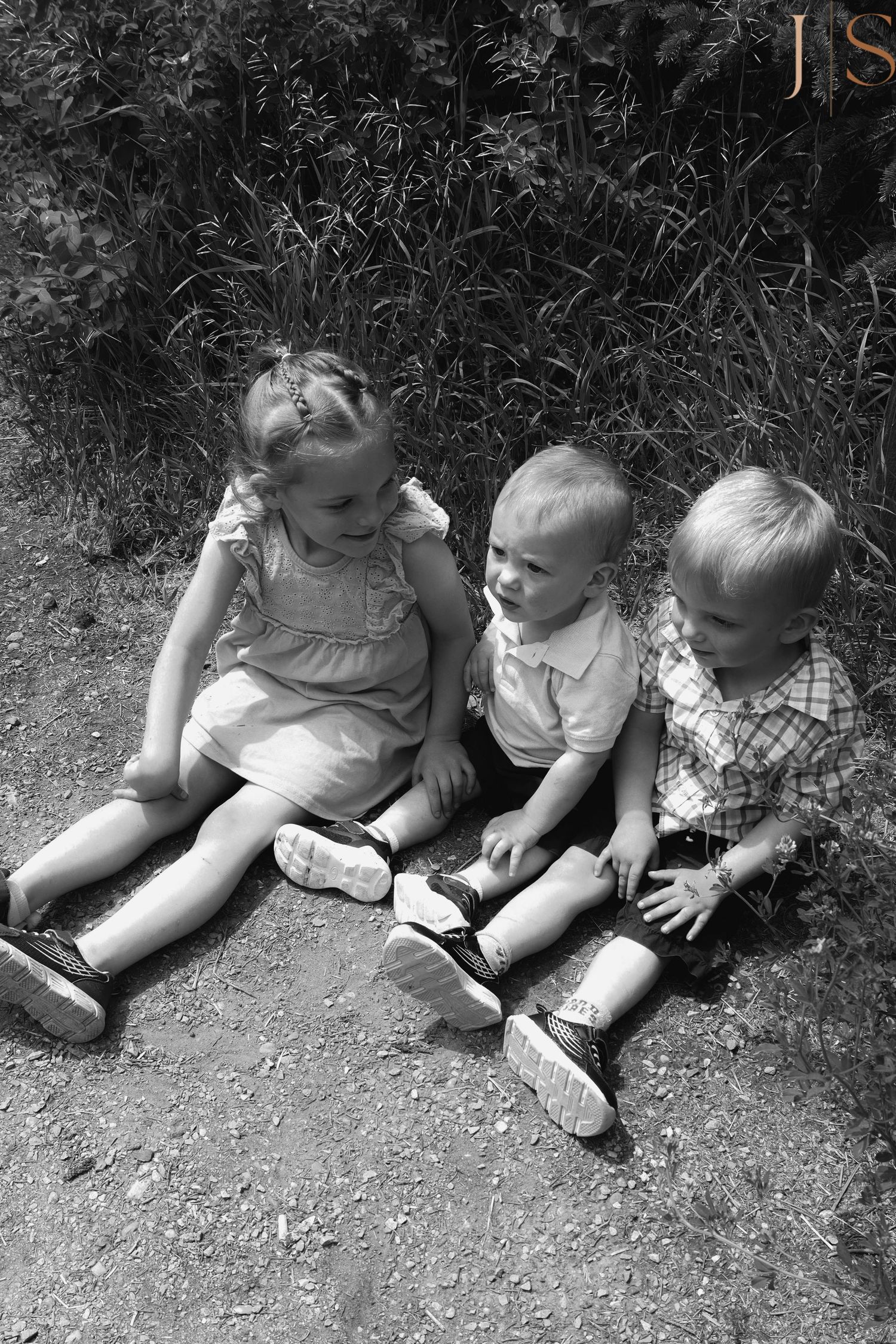 Three little children in vintage clothing lie in a row together on the ground in a black and white photograph series.