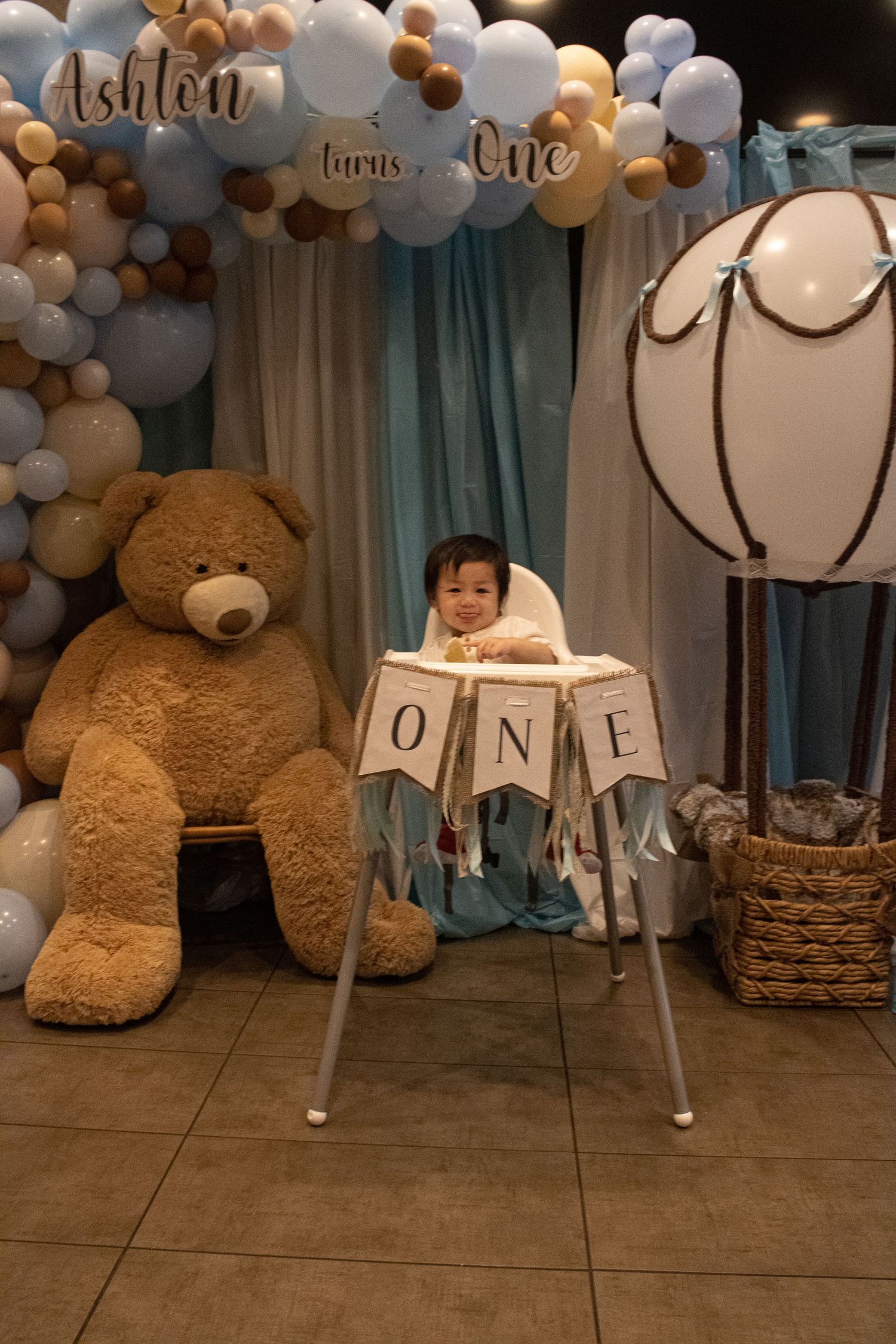 First birthday setup with large teddy bear, balloon arch, and baby in high chair.
