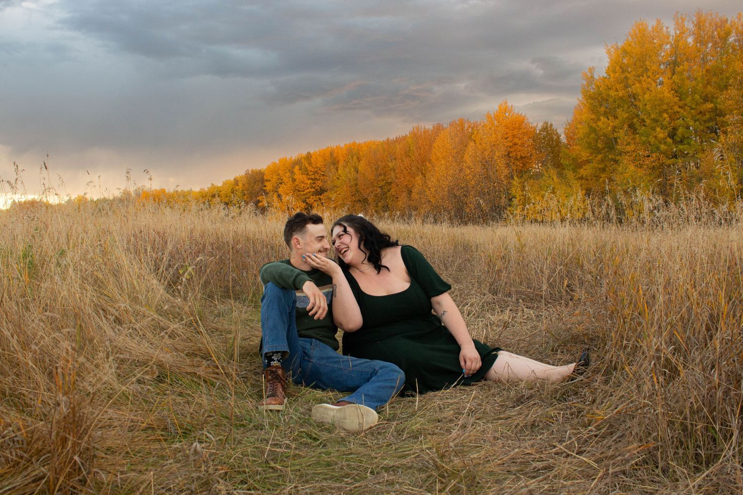 Couple sitting in golden autumn field at sunset with dramatic clouds overhead.