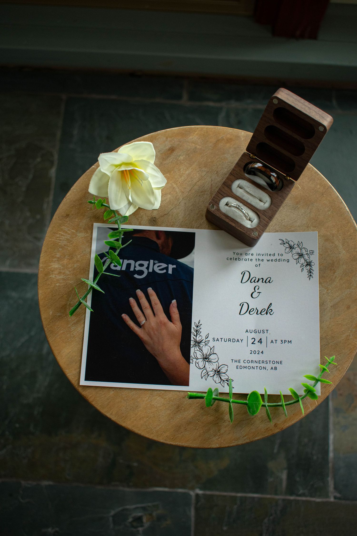 Wedding invitation and ring box displayed on wooden table with white rose and greenery decor.