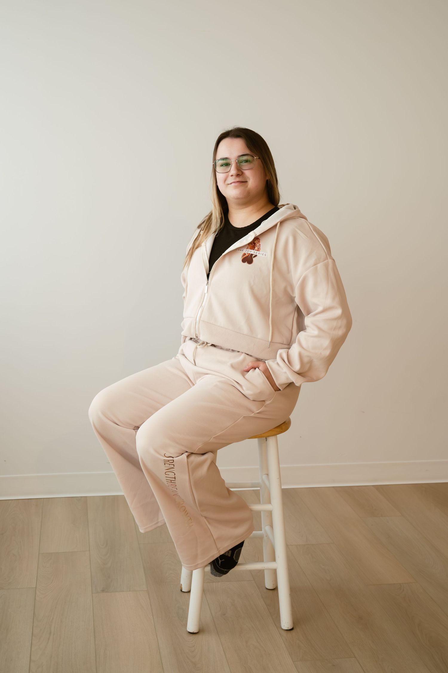 Young woman in pink tracksuit sitting on white stool, smiling, wearing green glasses, against neutral background.