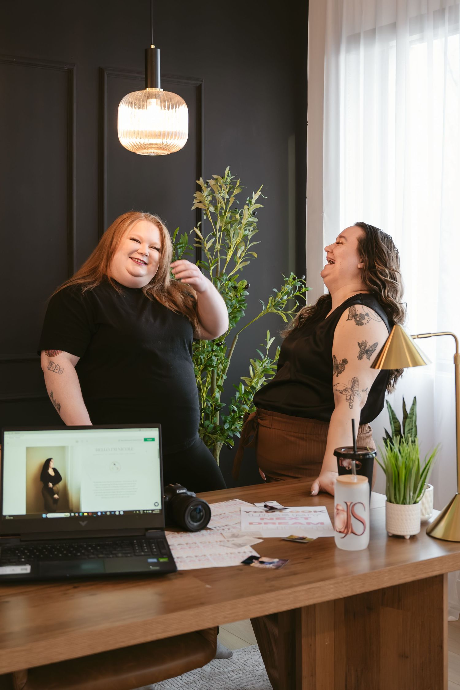 Two women laughing together at a wooden desk with a laptop, camera, and plants in a modern dark-walled office.