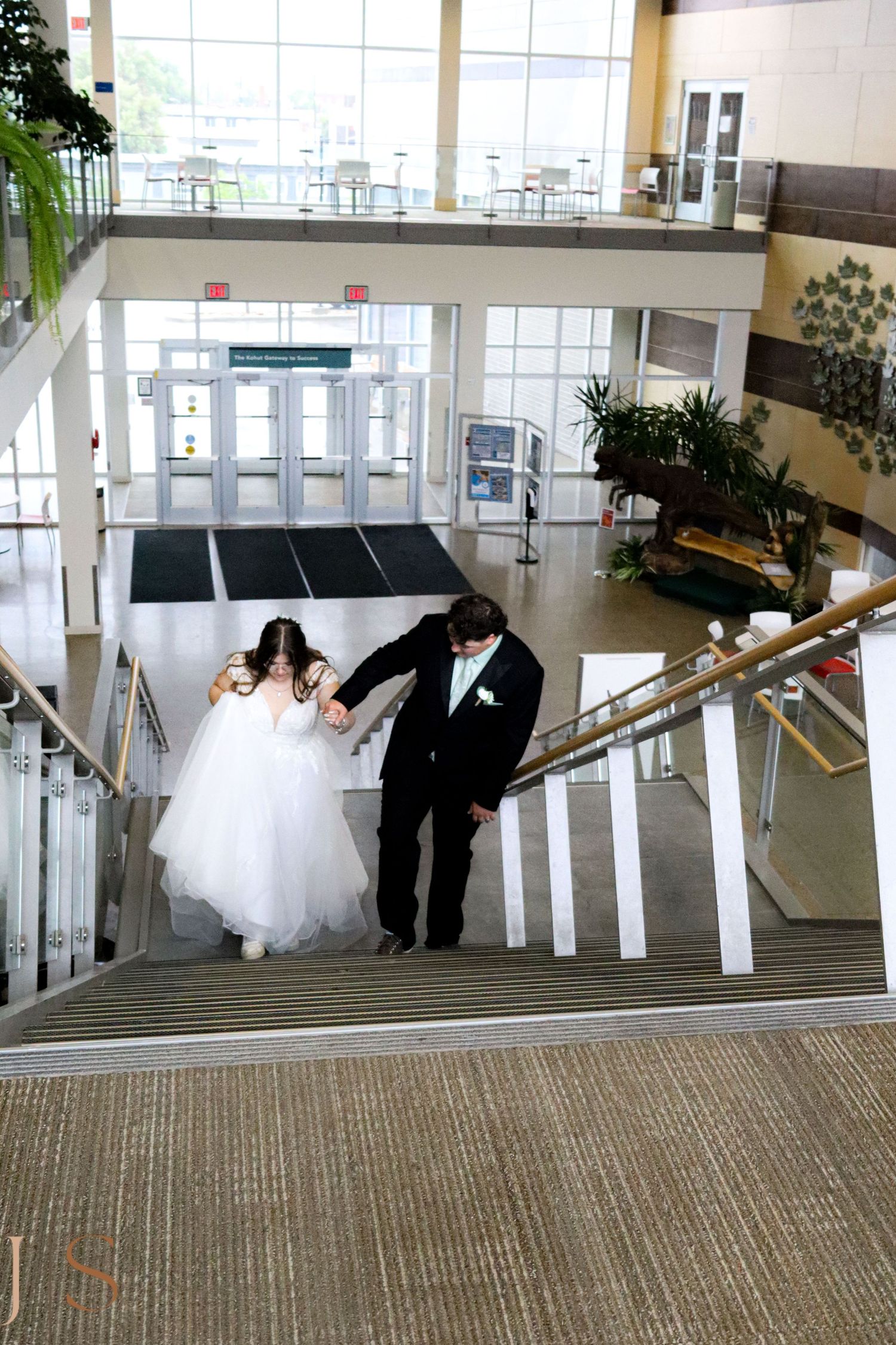 A bride in white dress and groom in tuxedo descend modern interior stairs together.