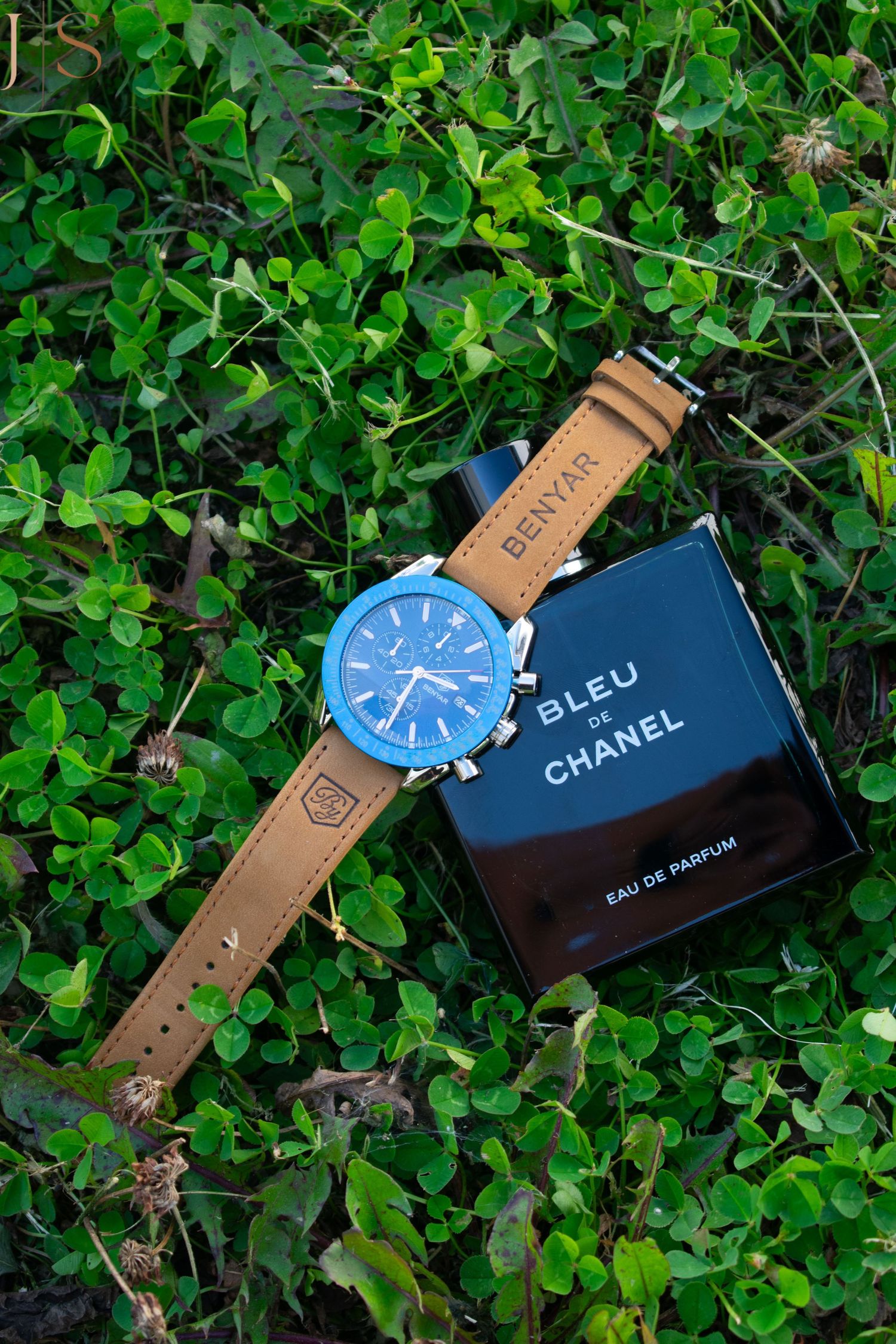 Stylish watch with brown leather strap and blue face displayed next to Bleu de Chanel perfume on green foliage background.