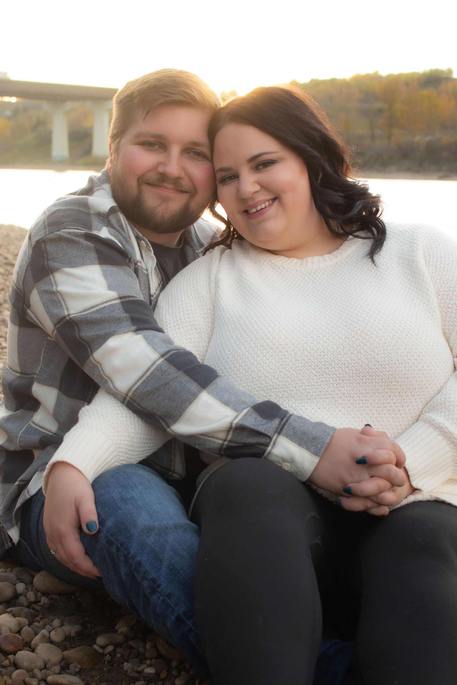 Couple sitting together on rocky shore at sunset in casual attire sharing tender moment.