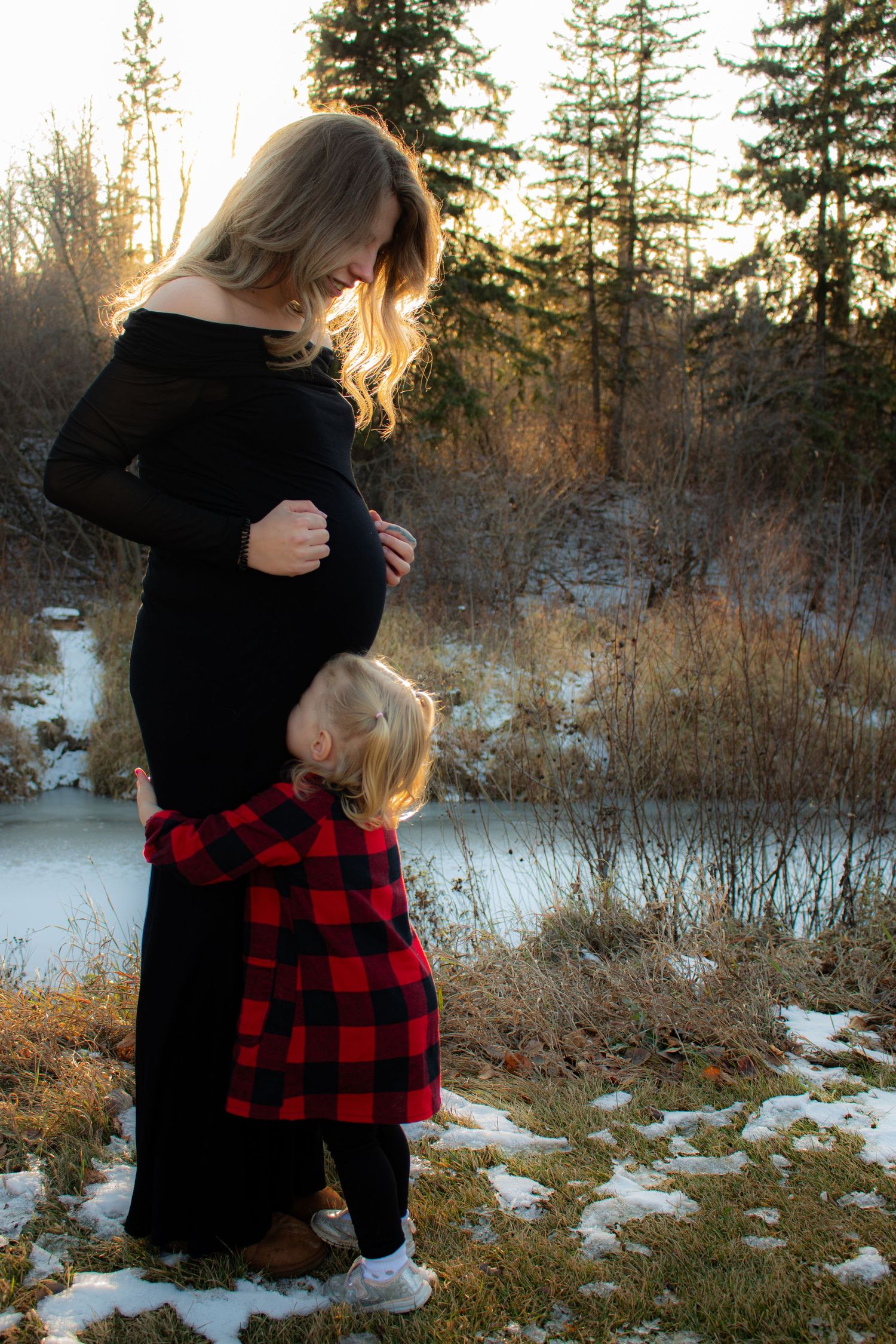 Winter maternity scene with black dress and red plaid accent against snowy landscape.