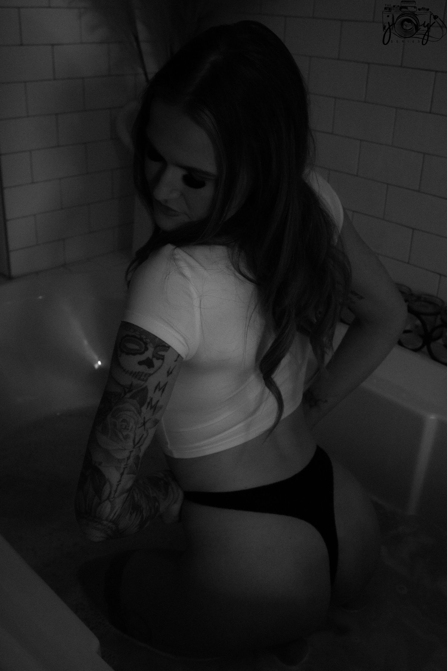 Black and white photo of person wearing white crop top with visible arm tattoo sitting in bathroom corner.