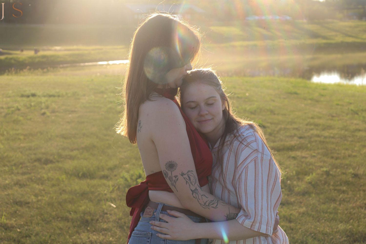Two figures share a tender embrace in a sunlit field at golden hour with a pond visible in the background.