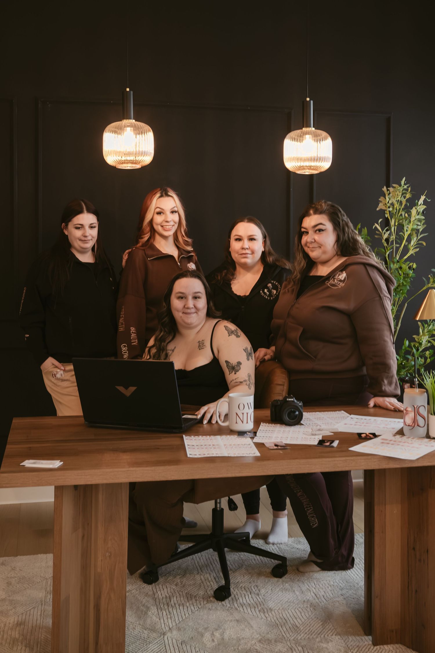 Four women pose together in a modern office with warm pendant lights, one seated at a wooden desk with a laptop.