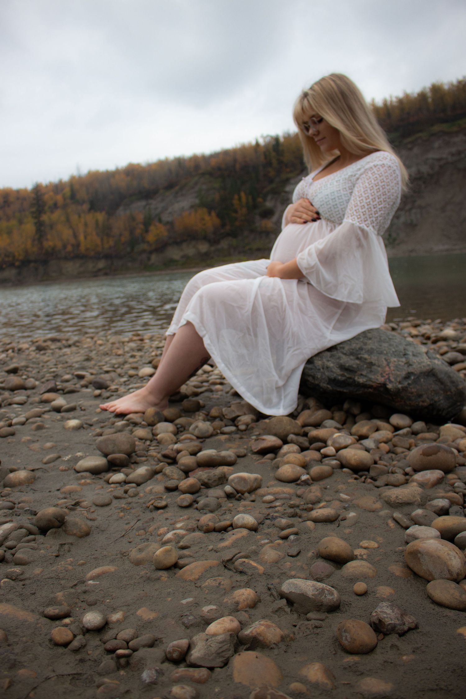 Maternity photo on rocky riverbank with woman in white dress during autumn.