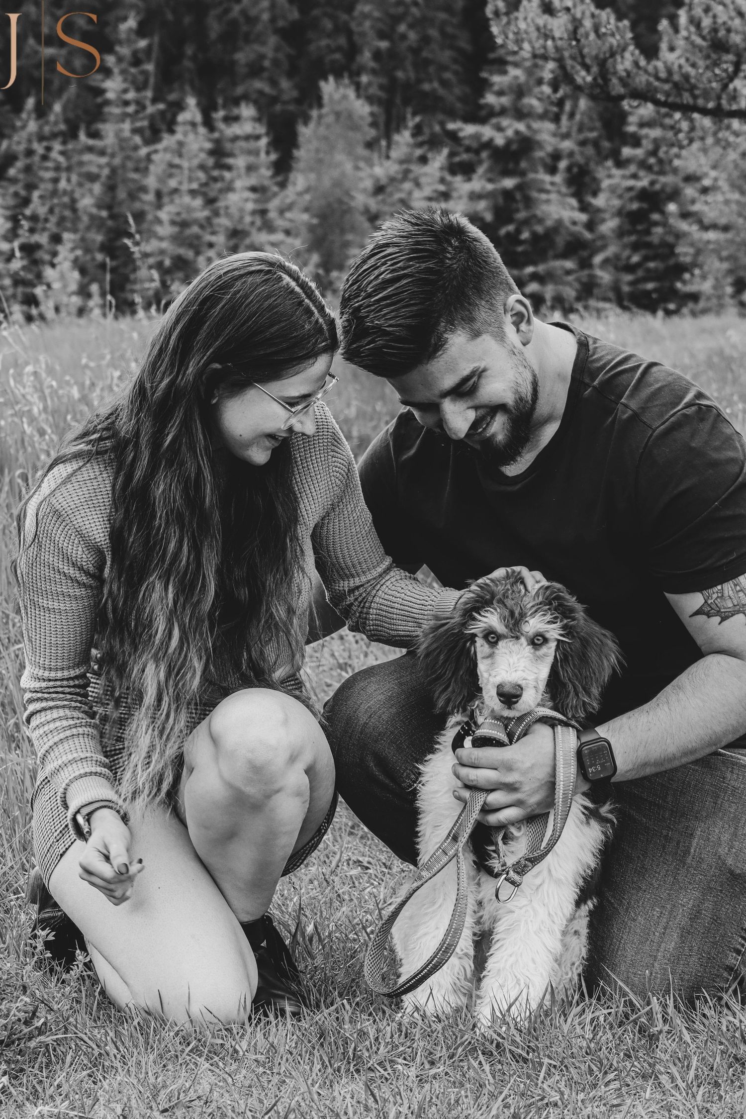 A couple lies in grass outdoors sharing tender moments with their husky dog in black and white photography.