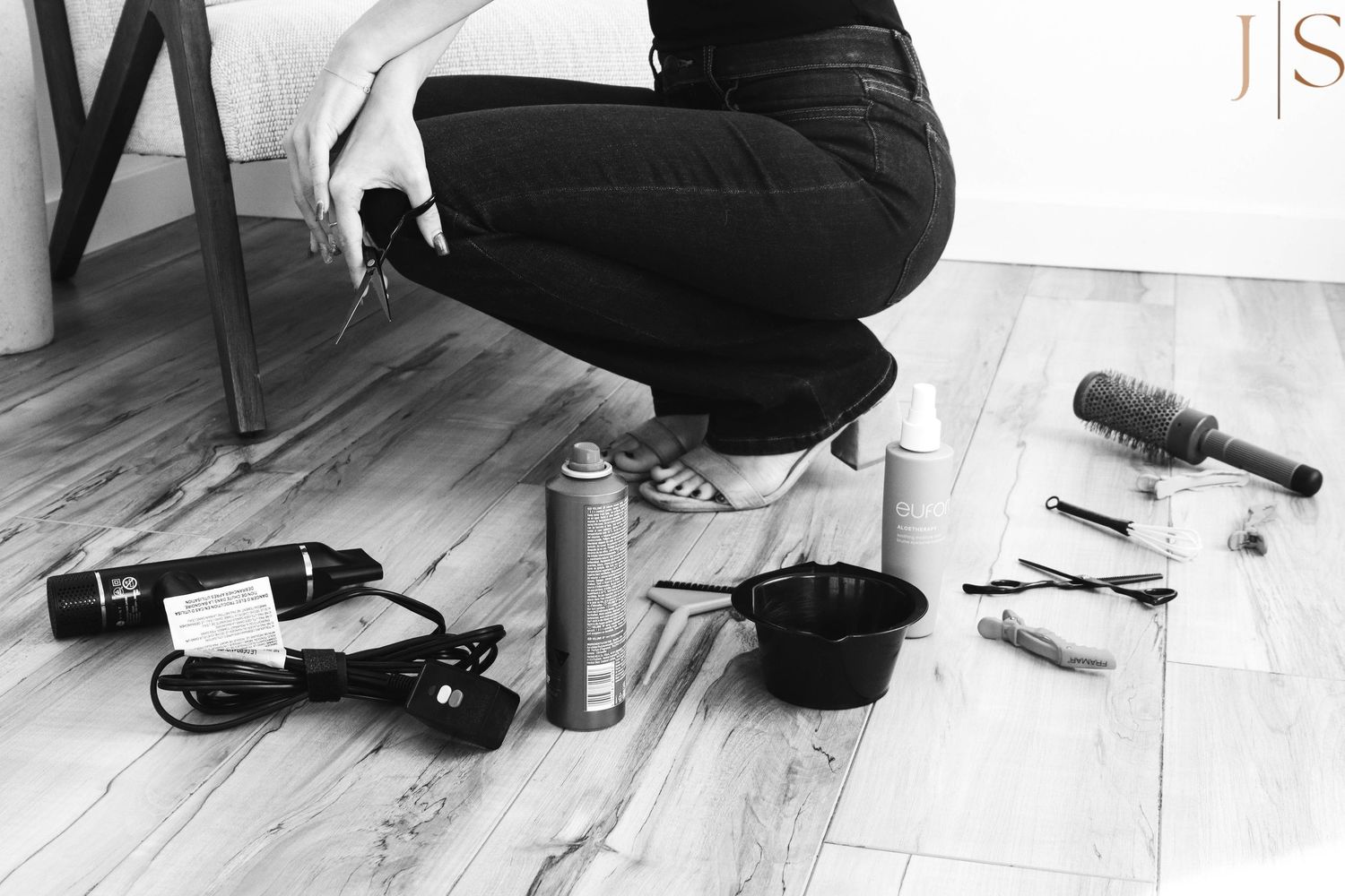 A person in black clothing kneels on wooden floor with various hair styling tools and products scattered around.