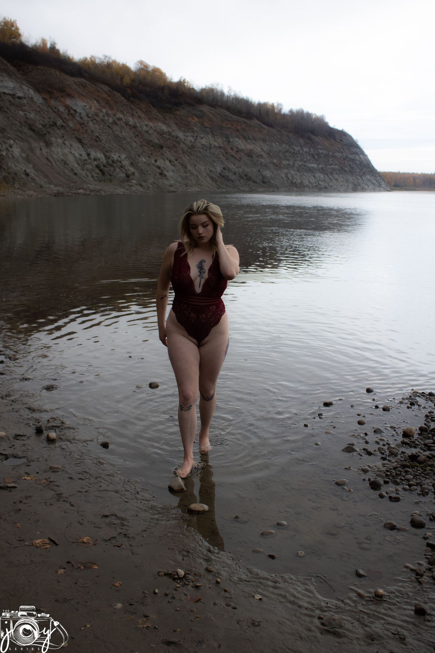 A person in a burgundy swimsuit poses at the edge of a lake with steep rocky cliffs in the background.