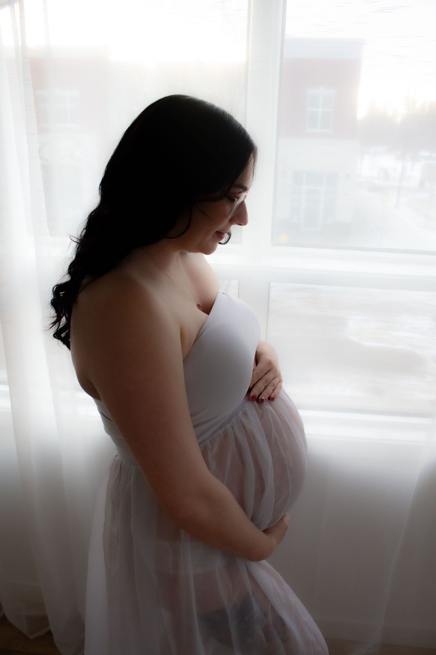 A maternity silhouette photo of an expectant mother in white dress by a window.