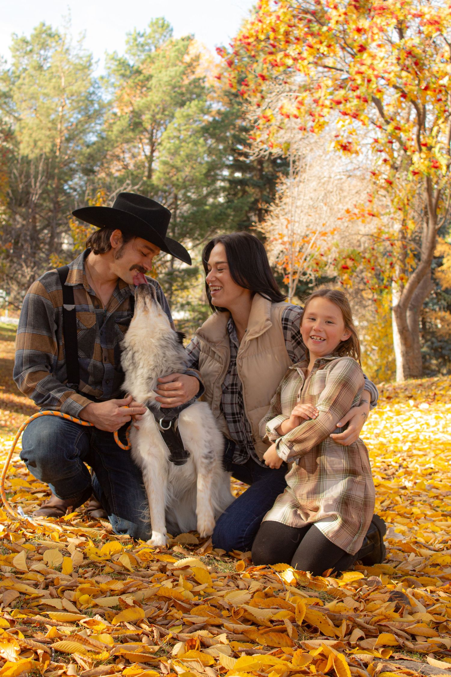 Family with pets poses together on fallen autumn leaves in woodland setting.