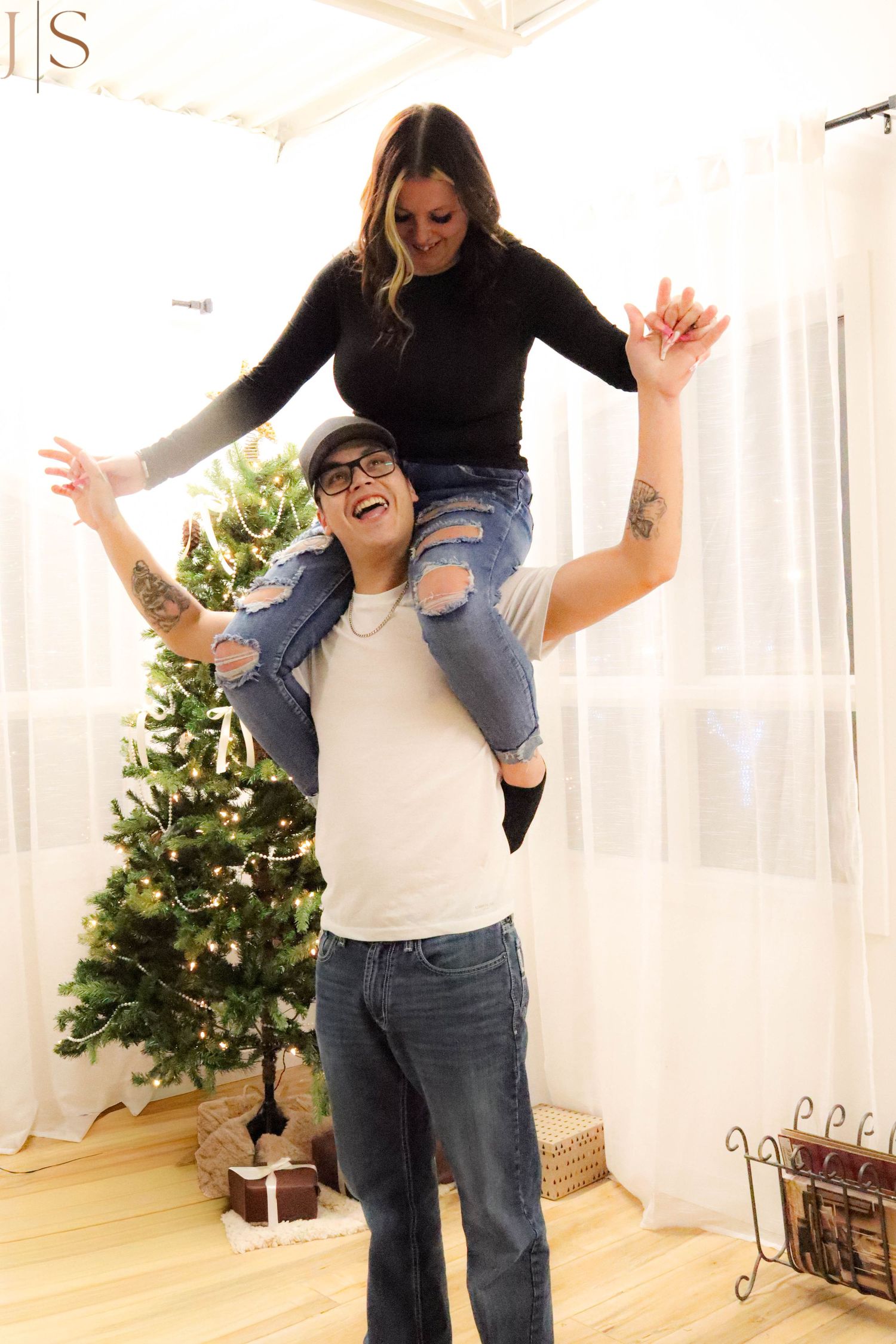 Two people playfully posing together in a bright room with a Christmas tree and holiday decorations in the background.