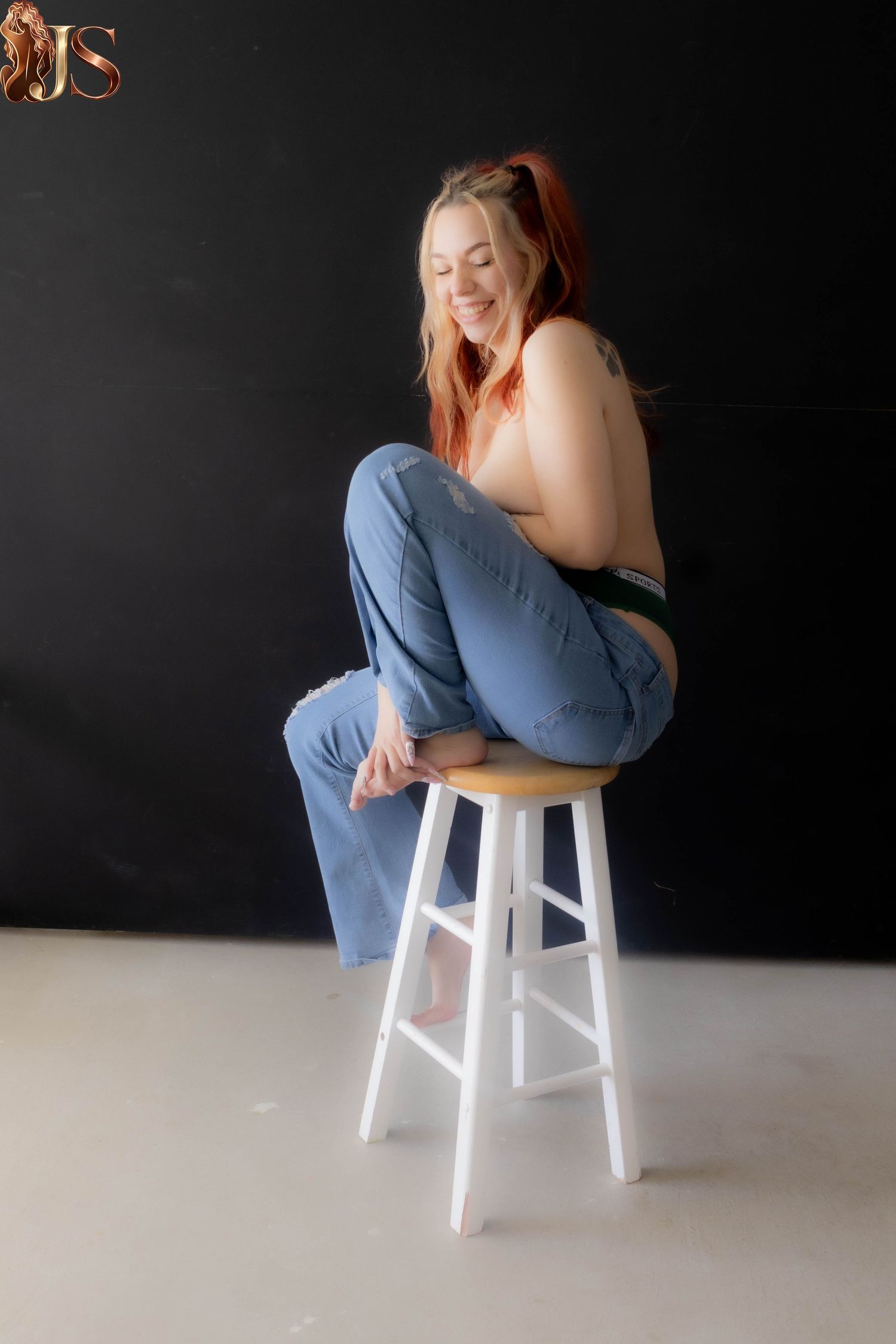 Figure sitting on white stool against dark background in casual denim clothing.