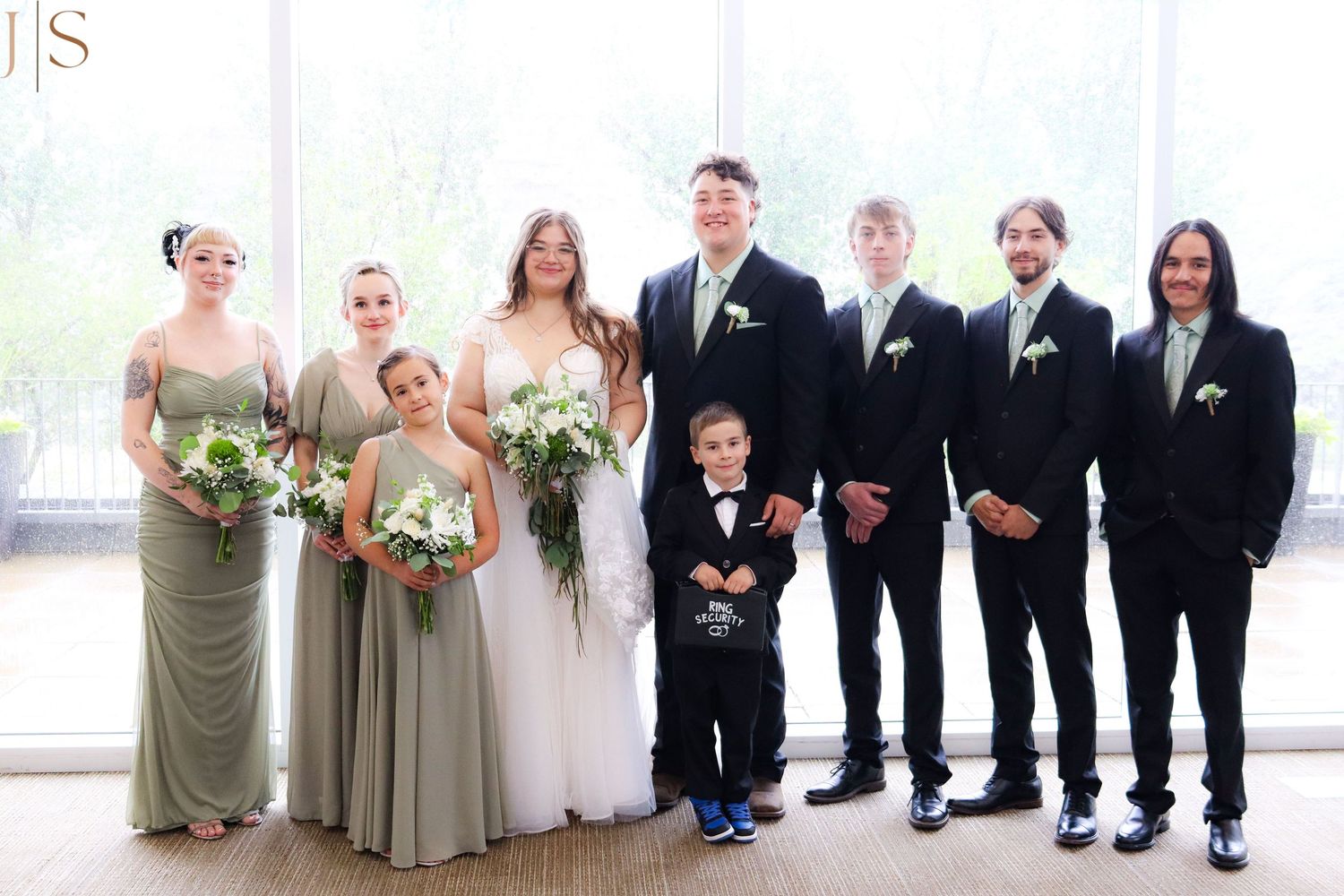 Wedding party members pose together in alternating black suits and sage green dresses for formal group photos.