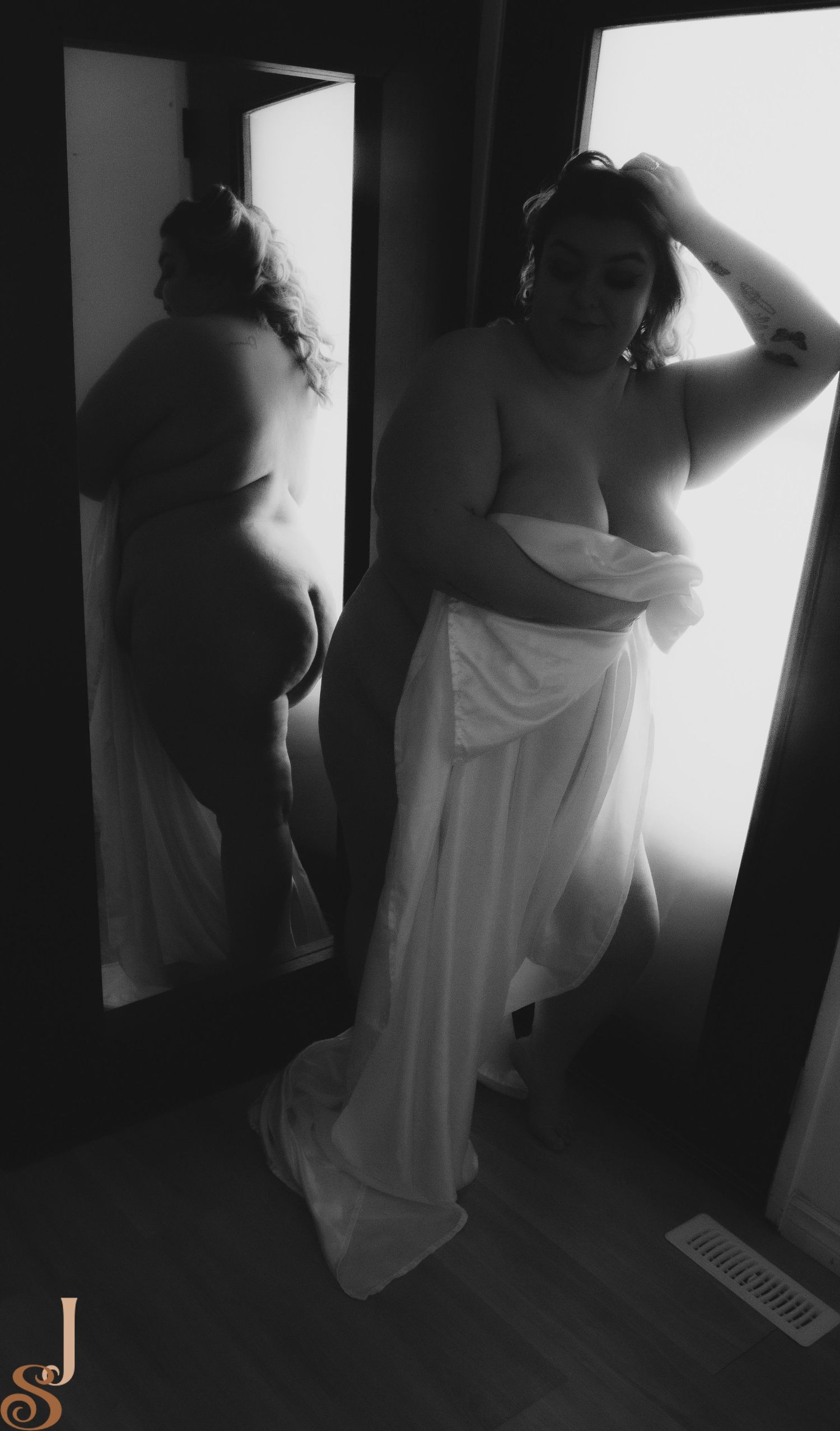 Black and white boudoir photo showing person in white flowing fabric standing beside mirror in dramatic lighting.