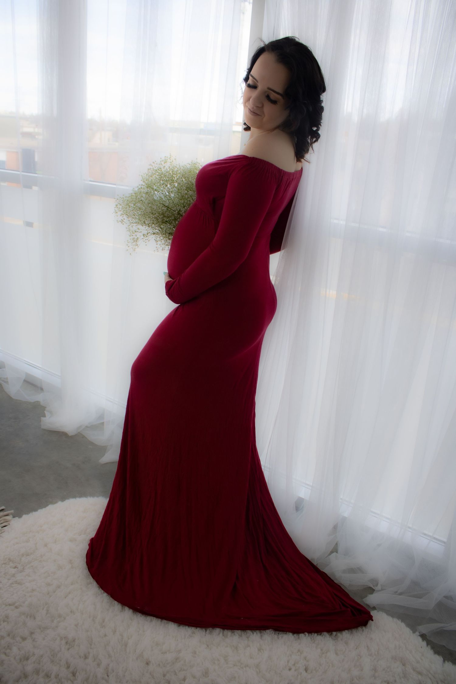 An elegant maternity portrait featuring a woman in a flowing red velvet dress holding baby's breath flowers.