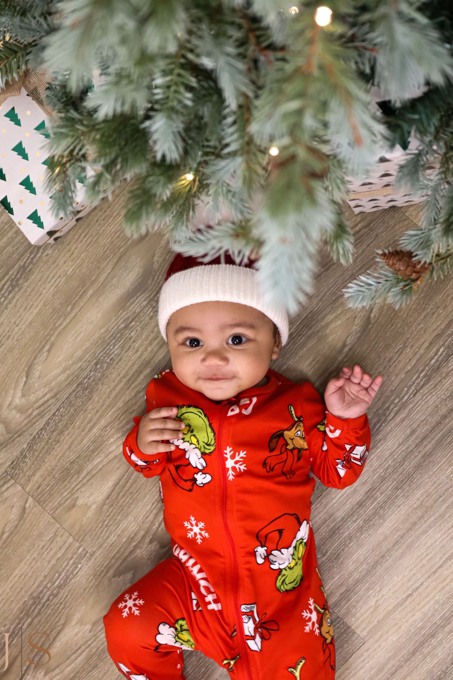 Baby wearing red holiday pajamas with festive prints lies under a decorated Christmas tree with twinkling lights.