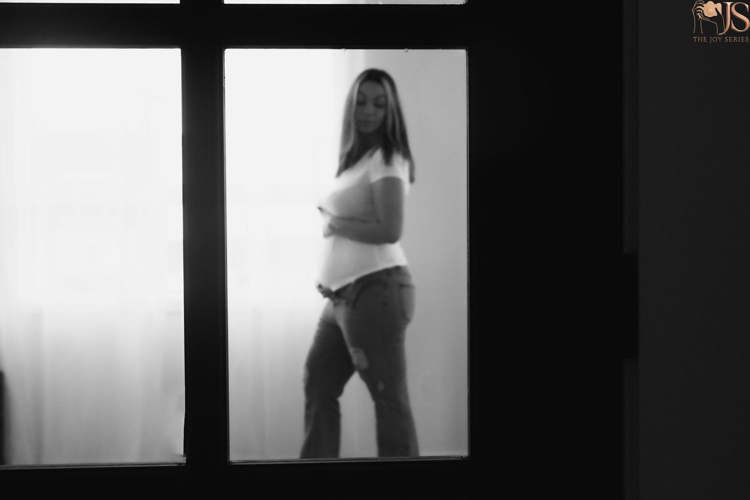 Black and white maternity photo of pregnant woman standing by window in white top and jeans.
