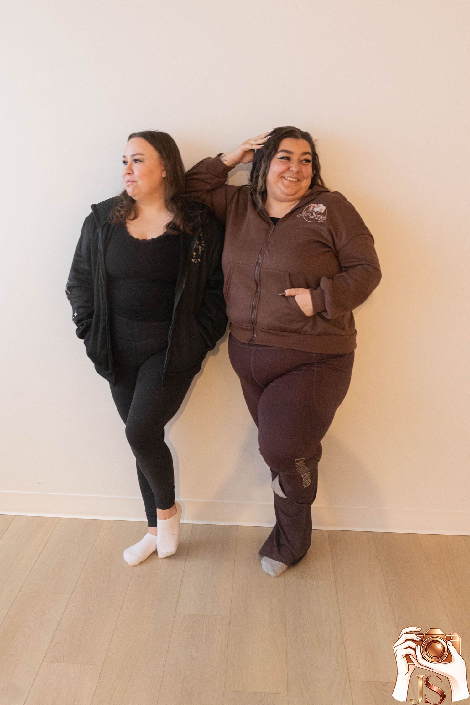 Two people in contrasting black and brown outfits strike playful poses against a white wall in a series of photos.