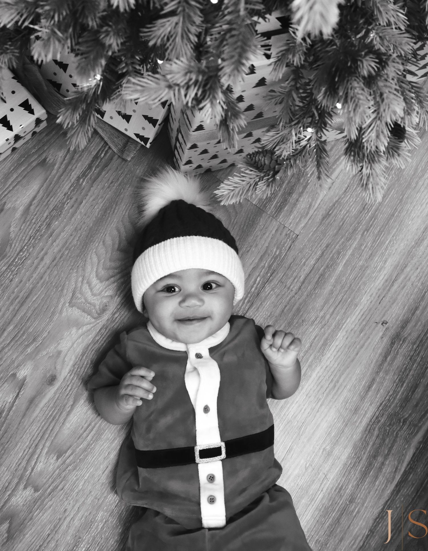 A series of black and white photos showing a baby wearing a white headband and dark clothing lying under a Christmas tree.