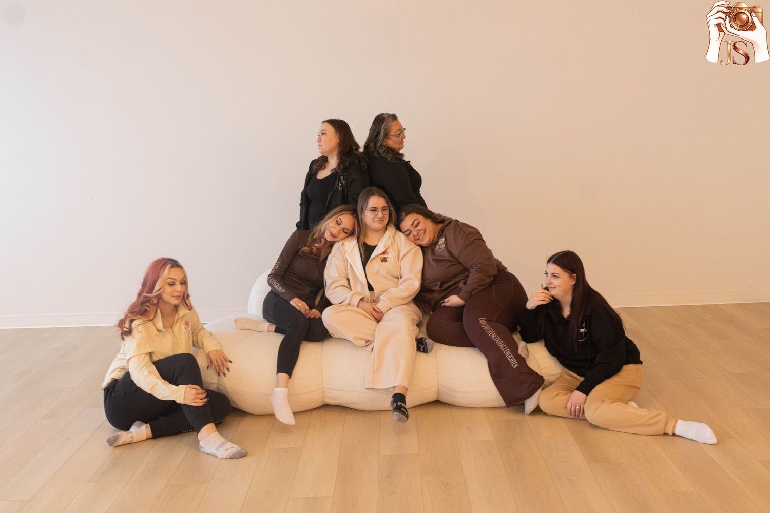 Group of people sitting and lying on floor in casual poses wearing light colored clothing in white studio space.