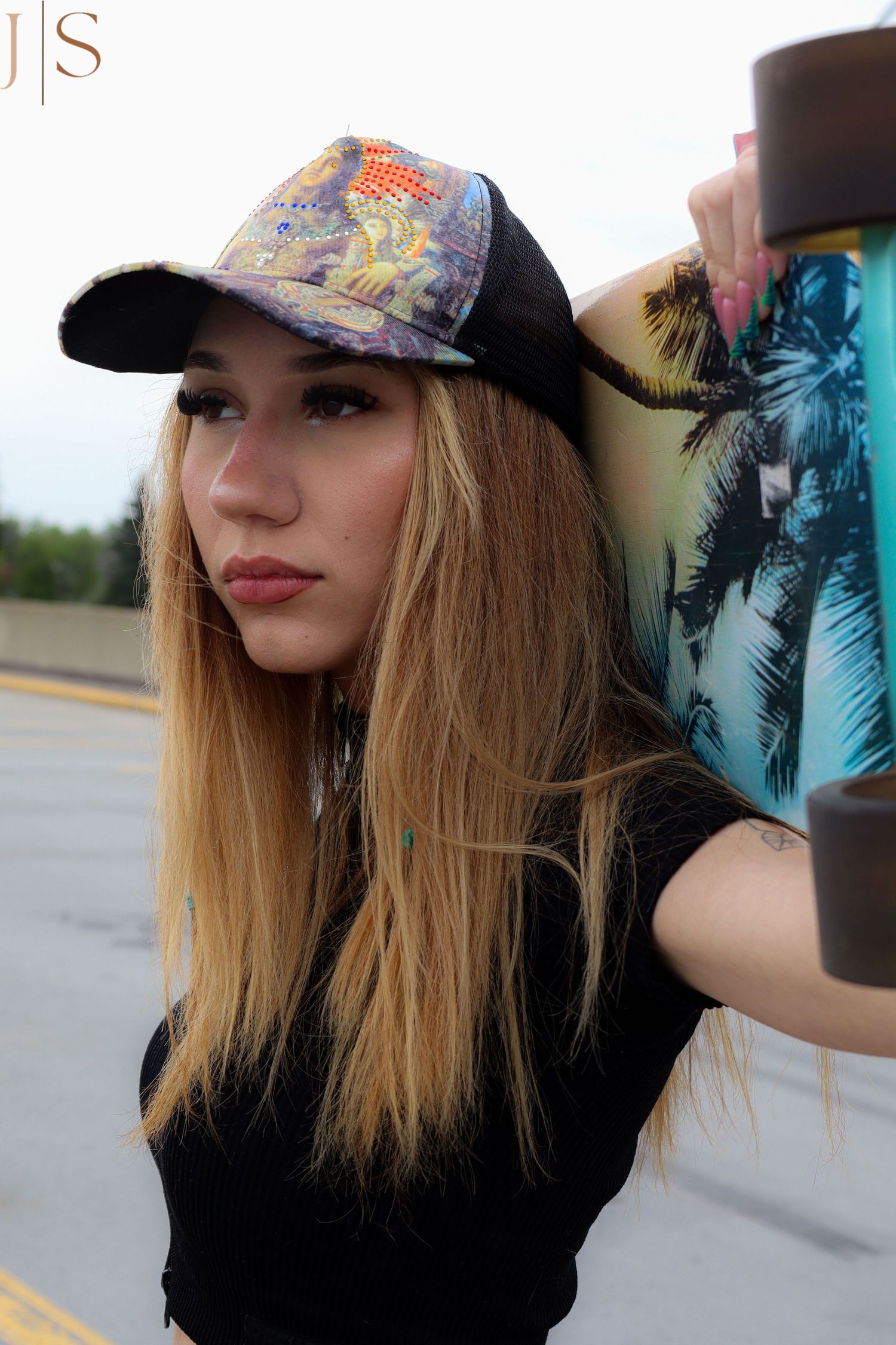 A person wearing a baseball cap with long blonde hair poses outdoors in casual attire.
