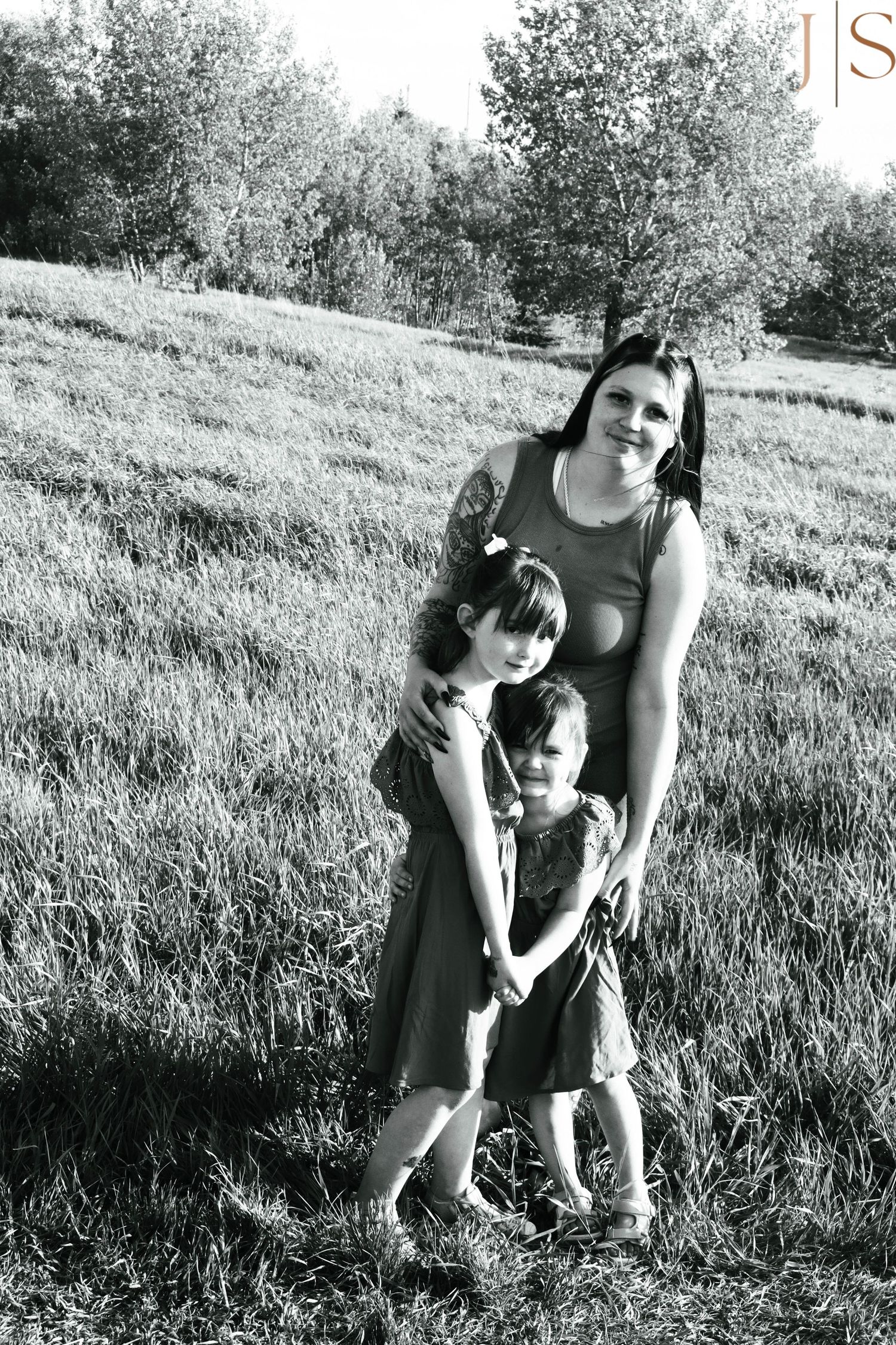 A black and white portrait of a family group embracing in a sunny meadow with trees in the background.
